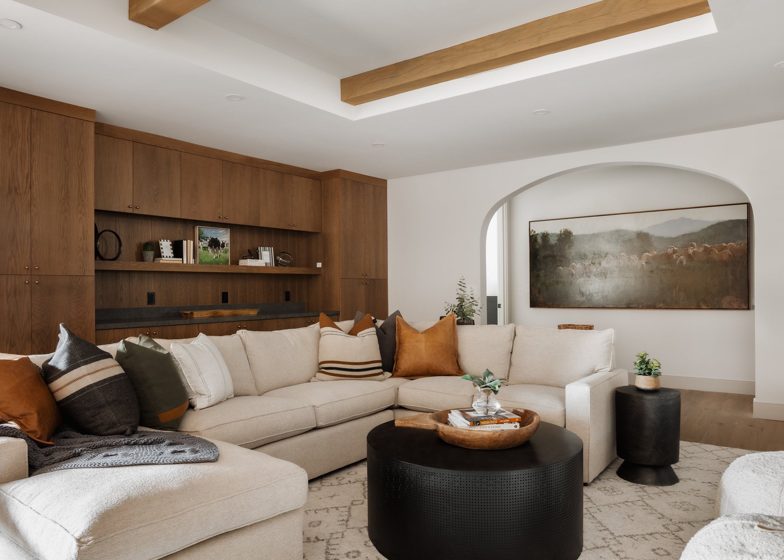 A modern living room with a large cream sectional sofa, decorative pillows, and a round black coffee table with a wooden bowl and books. The space features a built-in wooden wall unit, a large landscape painting, and small plants.