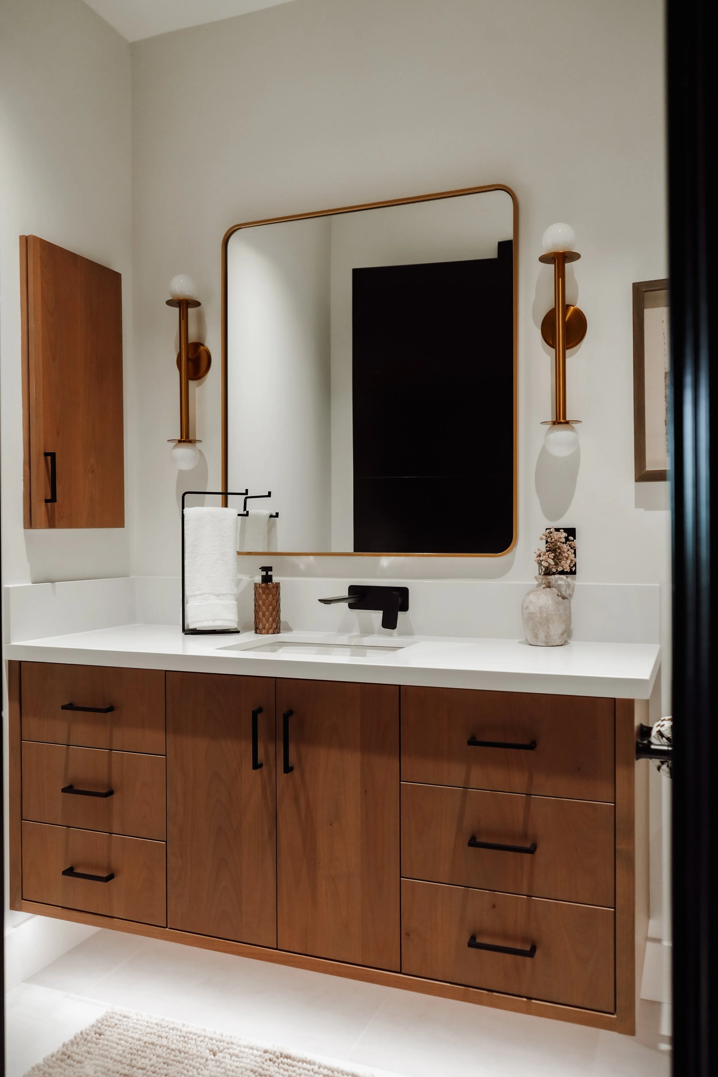 Modern bathroom vanity with wooden cabinets, white countertop, large square mirror, black faucet, and two wall-mounted lights with globe bulbs.