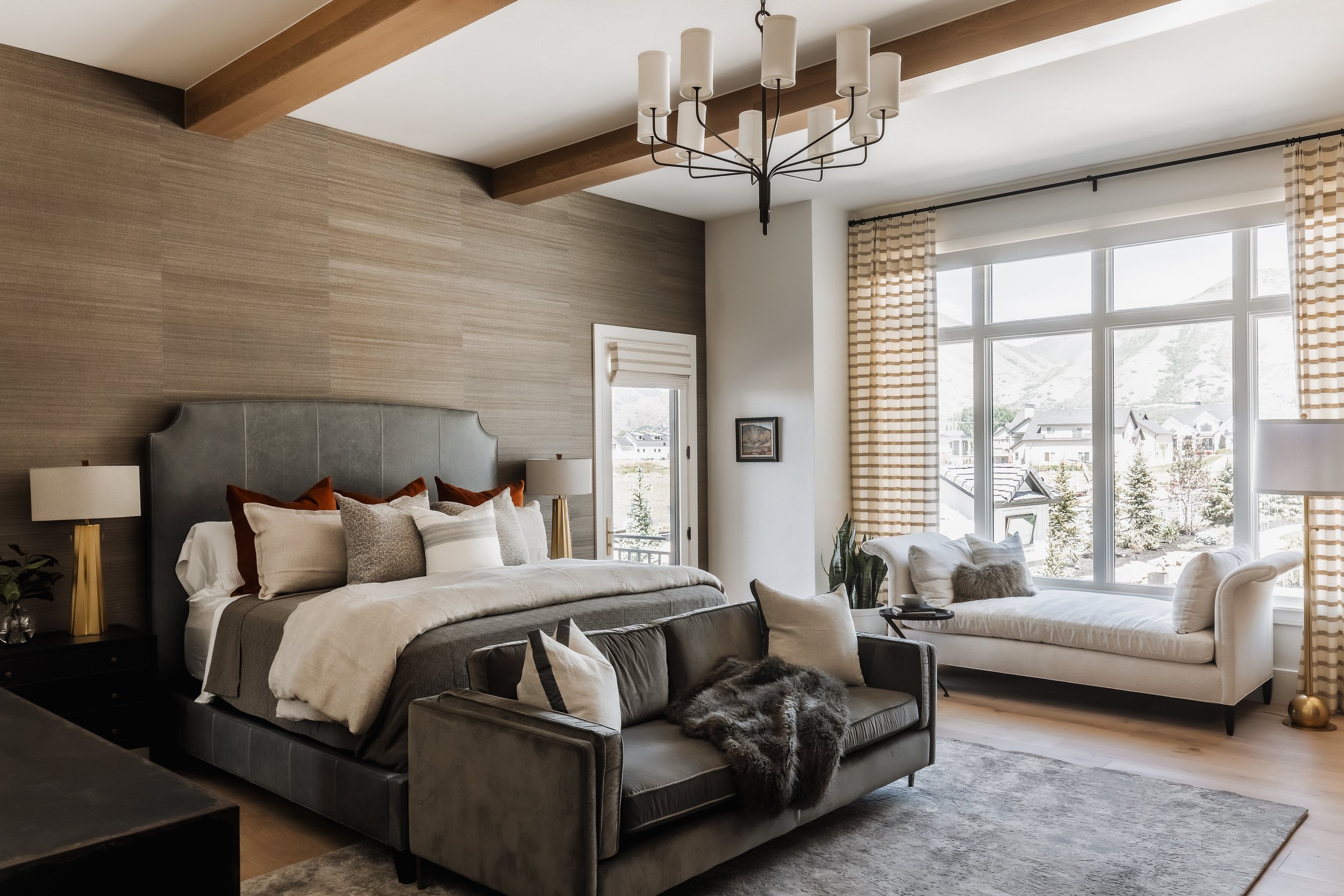 A bedroom with a large bed, a gray sofa, and a daybed by the window. Decor includes lamps, pillows, and a fur blanket. Large windows with curtains let in natural light and offer a mountain view.