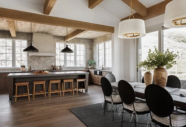 Modern kitchen and dining area with wooden beams, pendant lighting, stone backsplash, large windows, and a dining table with six black and white chairs.
