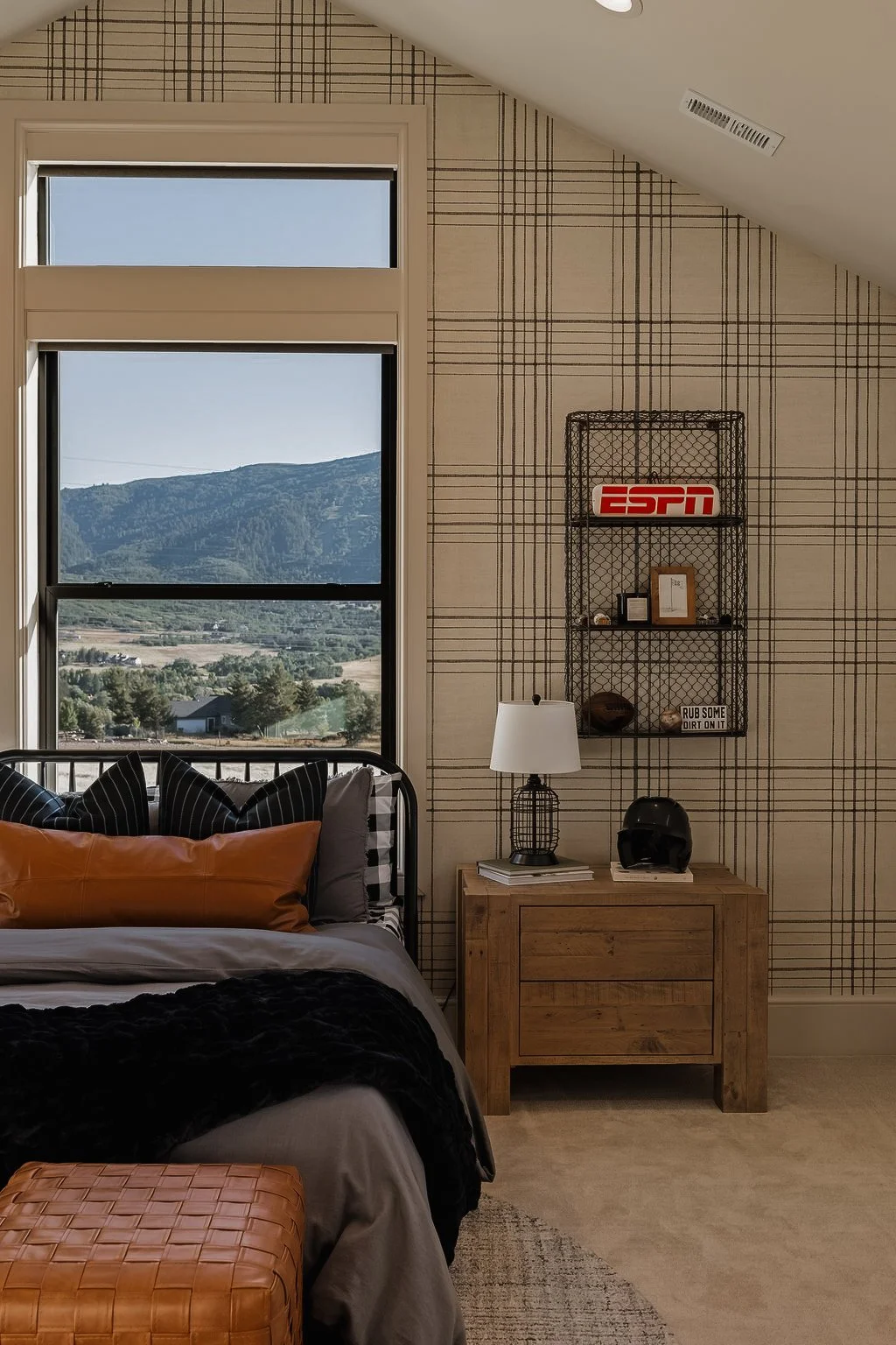 A bedroom with a large window showing mountain scenery, a bed with black and gray pillows and a tan pillow, a wooden nightstand with a white lamp and a black helmet, and a wall shelf with an ESPN sign and various decorations.