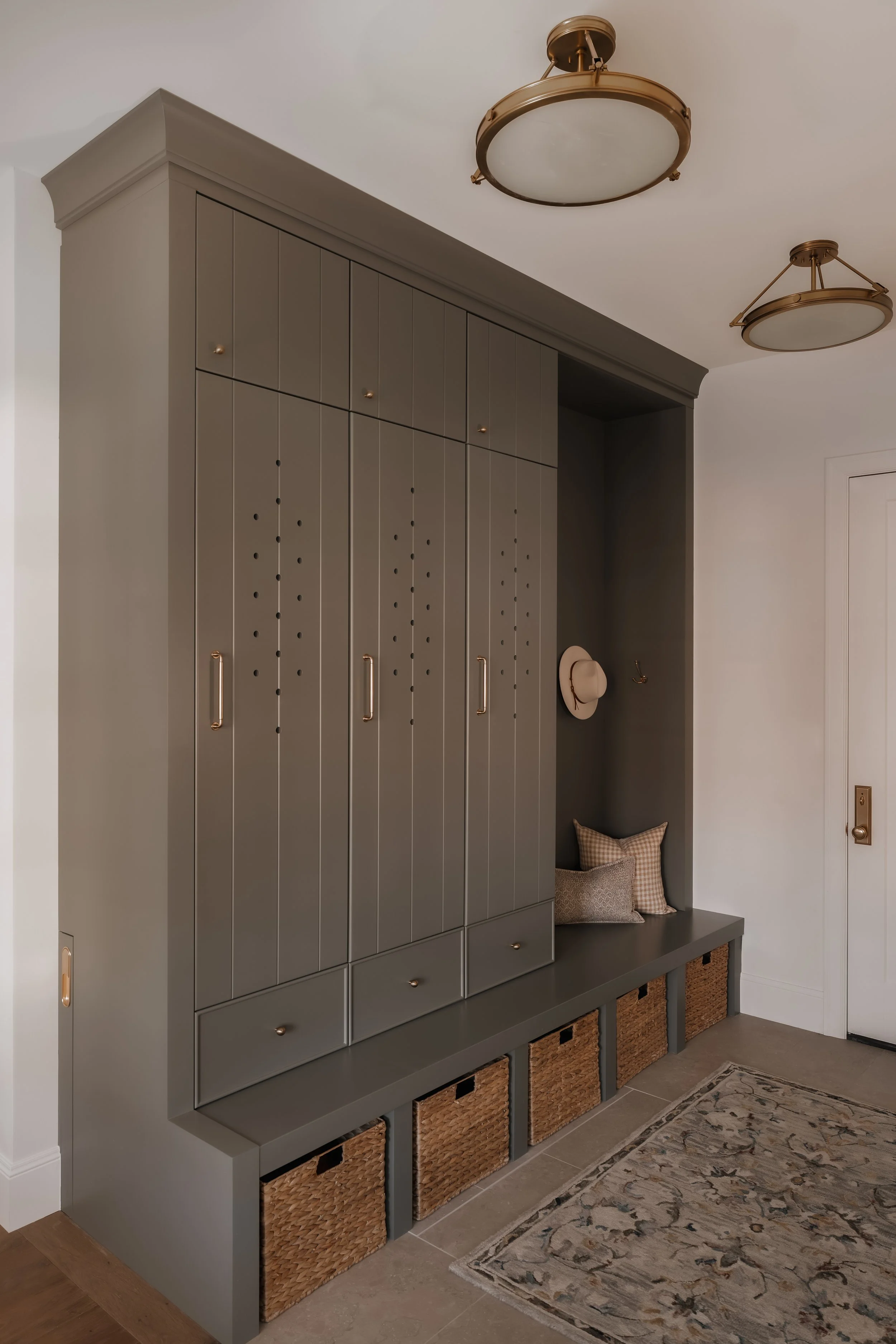 A modern gray built-in storage cabinet with drawers and baskets, a cushioned bench with pillows, a hat hanging on the wall, and ceiling lights in a home's entryway.