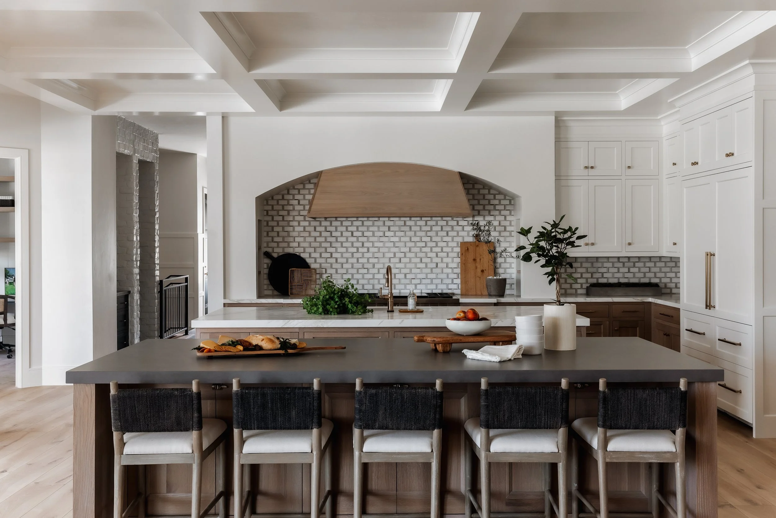 Modern kitchen with white cabinetry, a large island with a dark countertop, and decorative ceiling details. Sink, potted plant, and food items on the island. Brick backsplash and wooden accents.