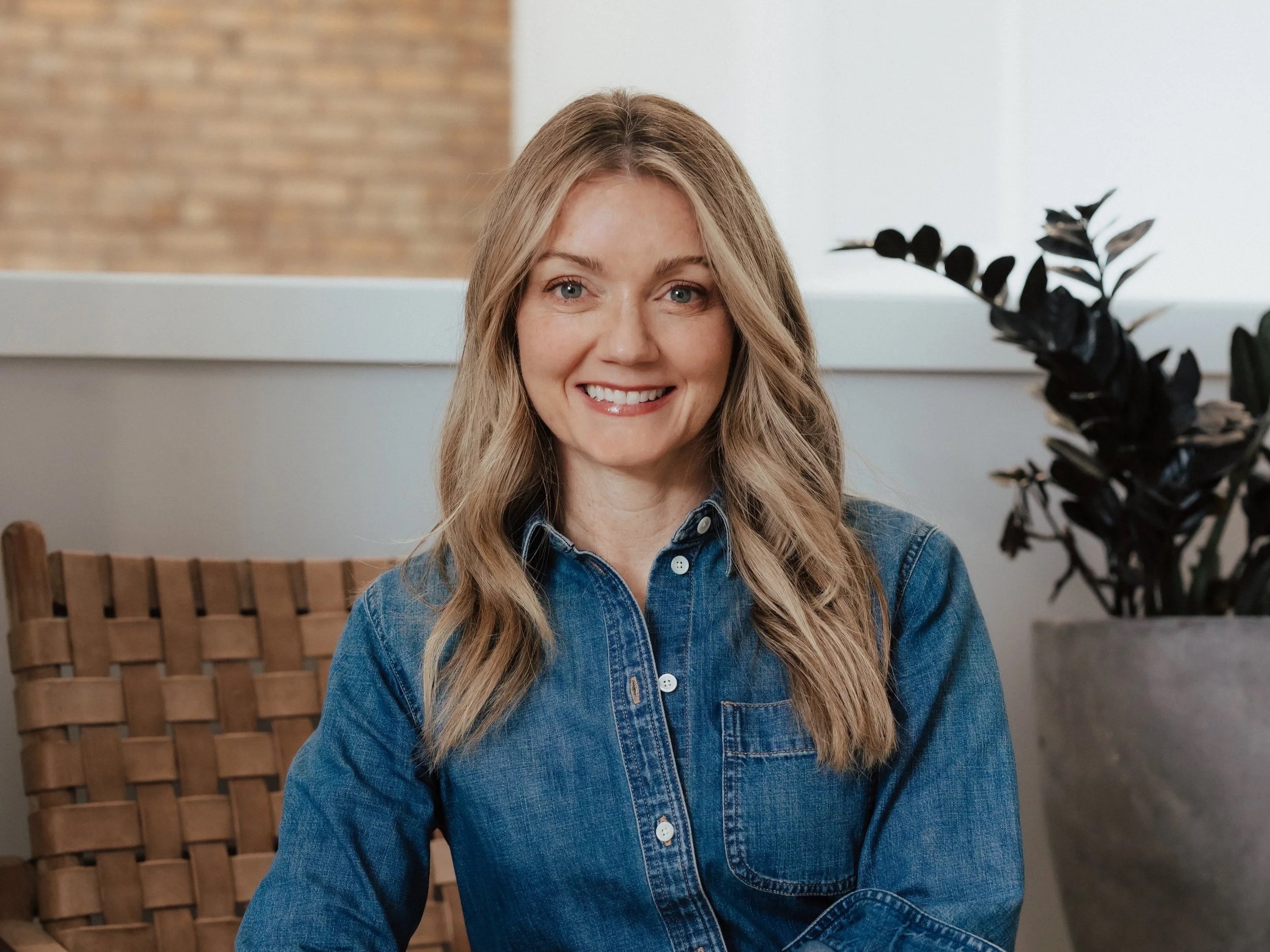 Woman with light brown wavy hair smiling, wearing a denim shirt, sitting in a cozy indoor space with a wooden chair and a large potted plant.