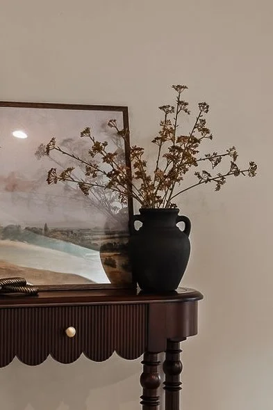 Dried branches in a black vase on a wooden table with a landscape picture in the background.
