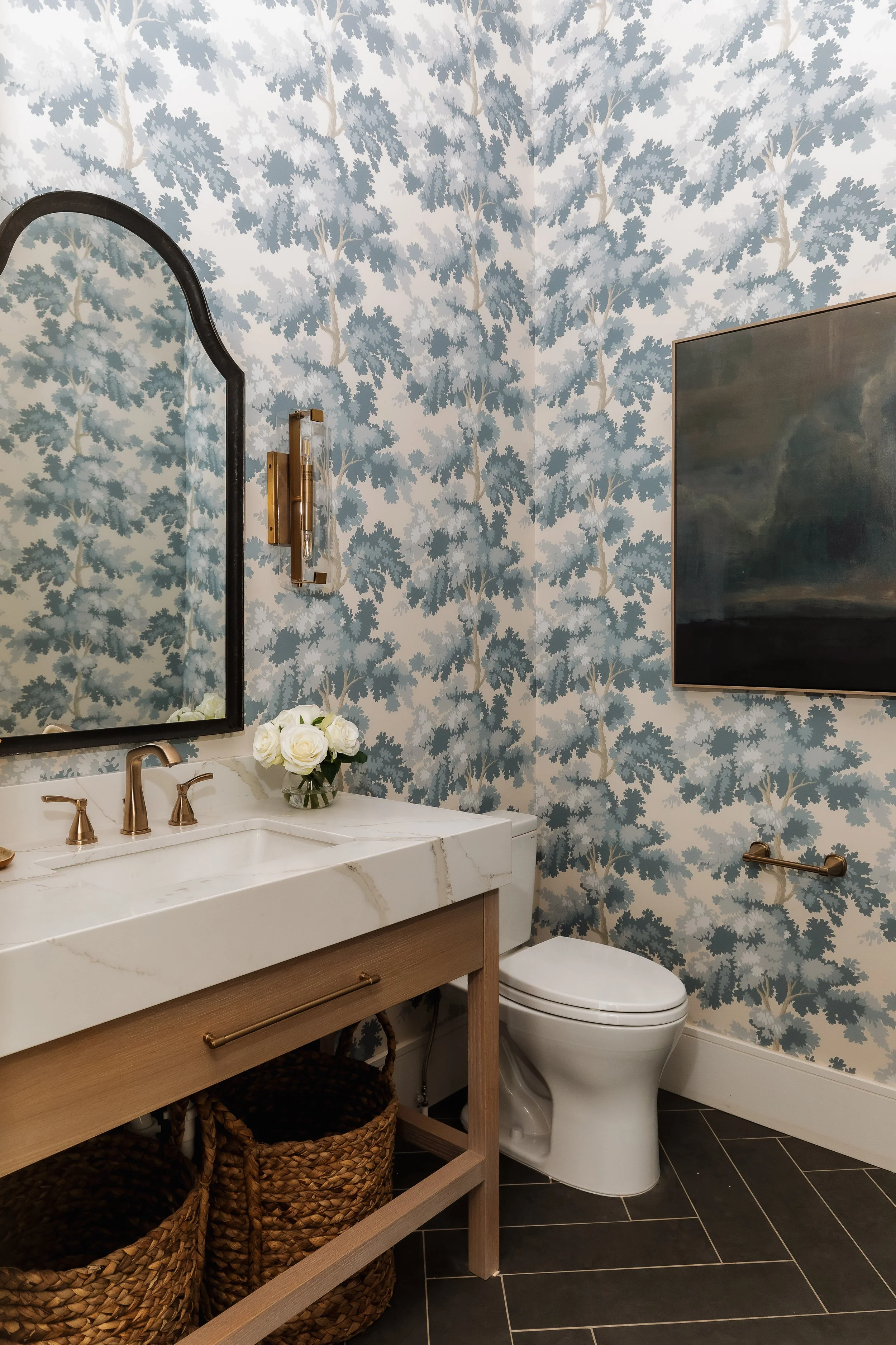 A bathroom with blue and white leafy wallpaper, a mirror, a faucet, white flowers in a glass vase, a wooden vanity with baskets underneath, a toilet, a framed picture, and a towel rail.