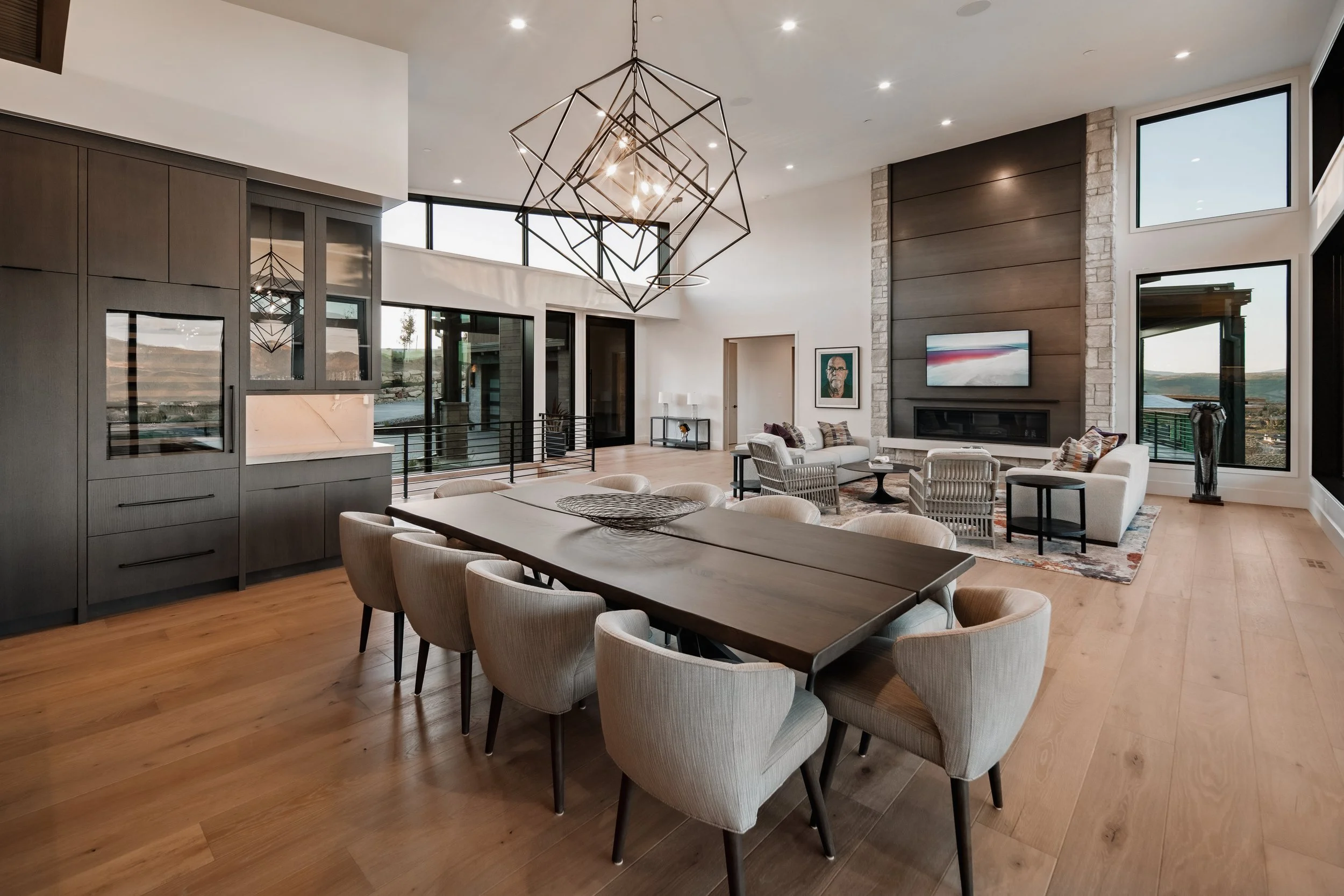 Modern open-concept living and dining room with large windows, a wooden dining table surrounded by beige chairs, a cozy sitting area with white sofas, armchairs, a fireplace, and contemporary art and lighting fixtures.