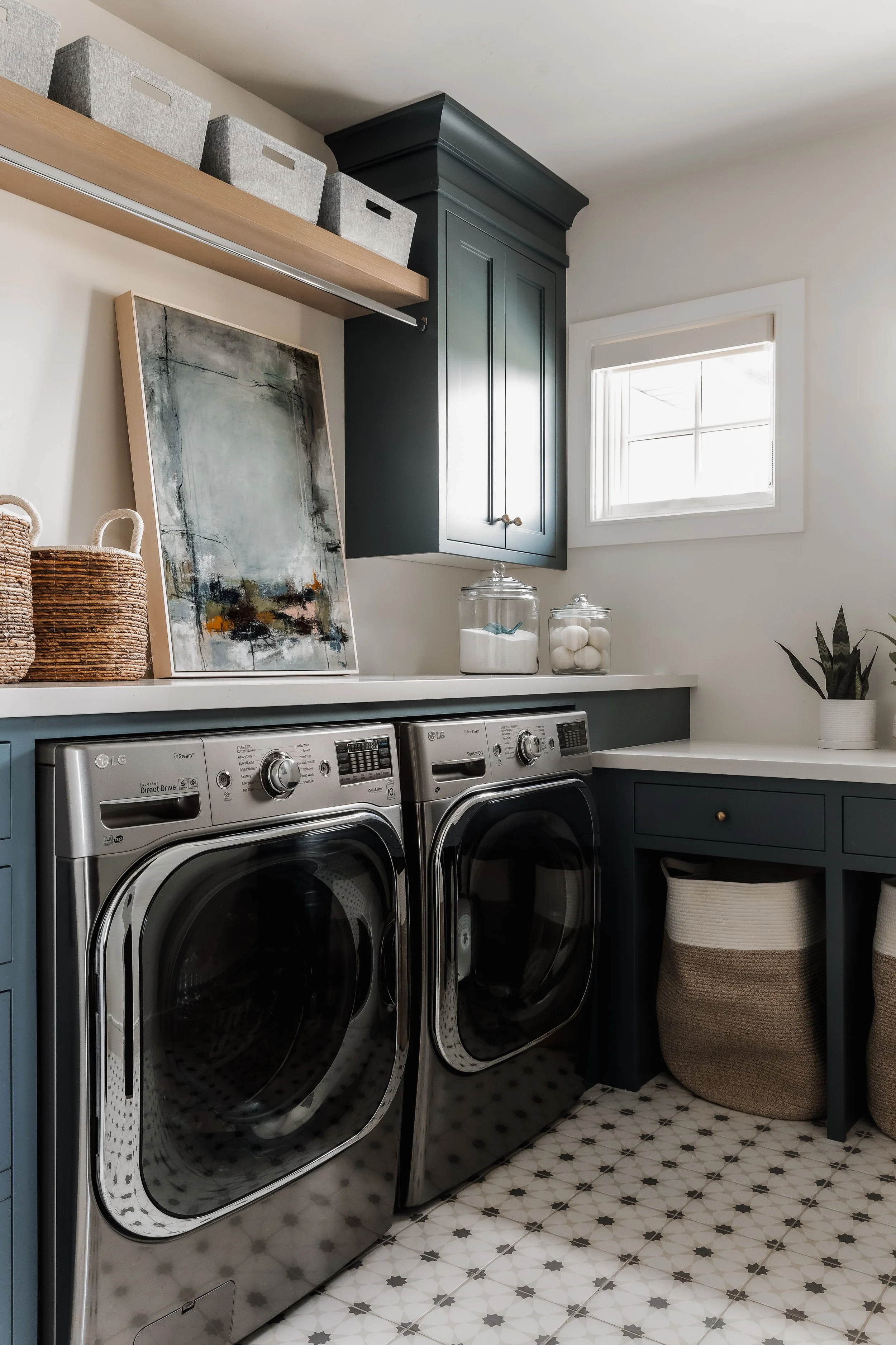 Modern laundry room with front-loading washer and dryer, dark blue cabinets, a small window, and decorative storage baskets.