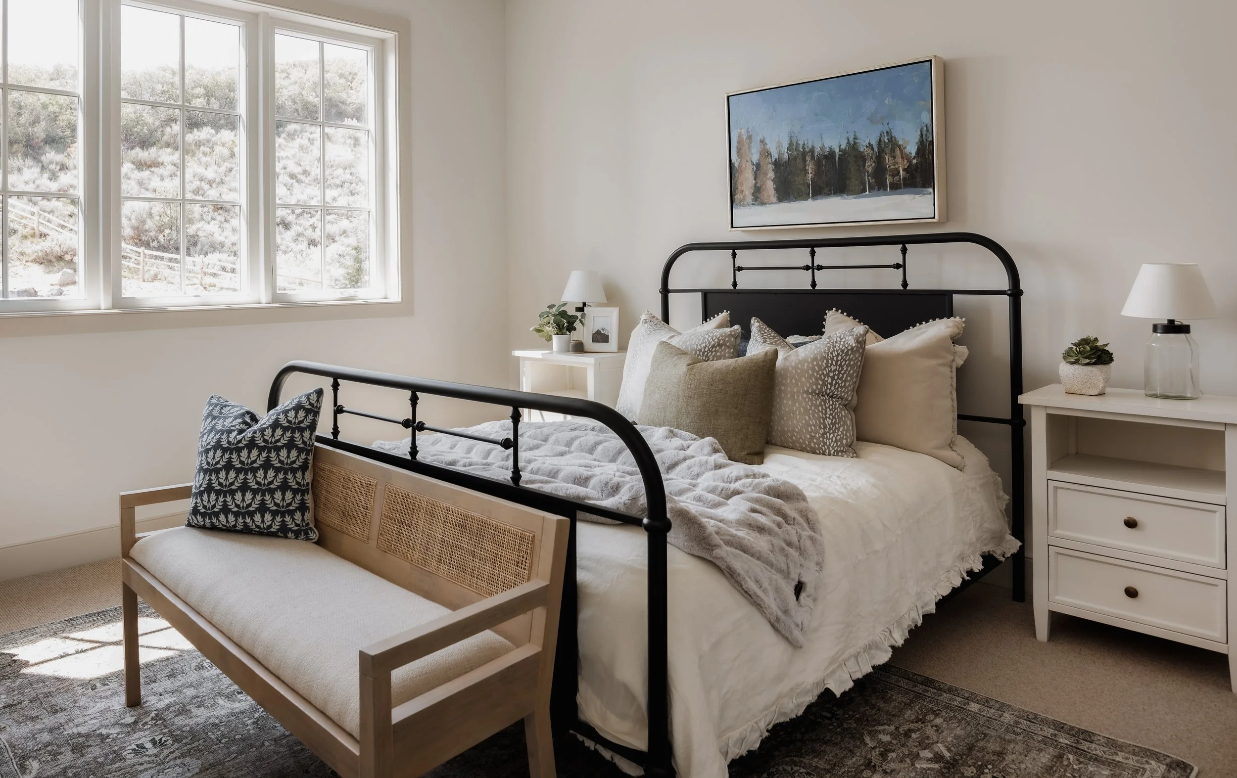 Cozy bedroom with large window, black metal bed frame, white bedding, gray and beige pillows, white nightstands with lamps and plants, and a landscape painting above the bed.