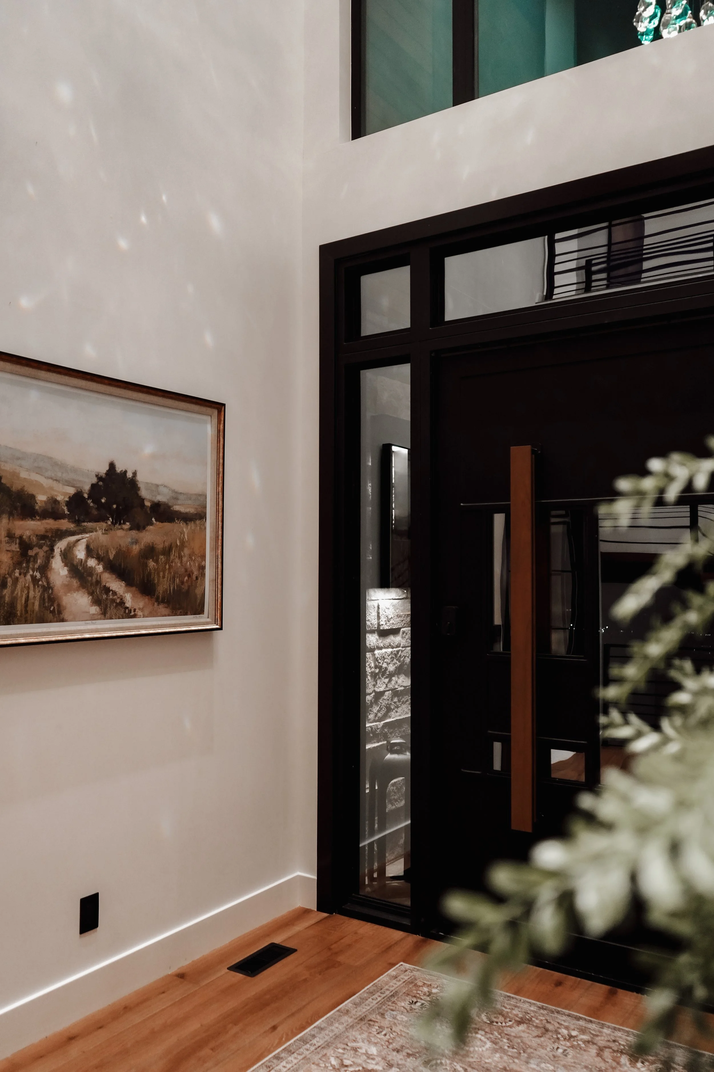 View of a modern entryway with a black door, a window, a painting of a countryside road, and a partially visible plant in the foreground.