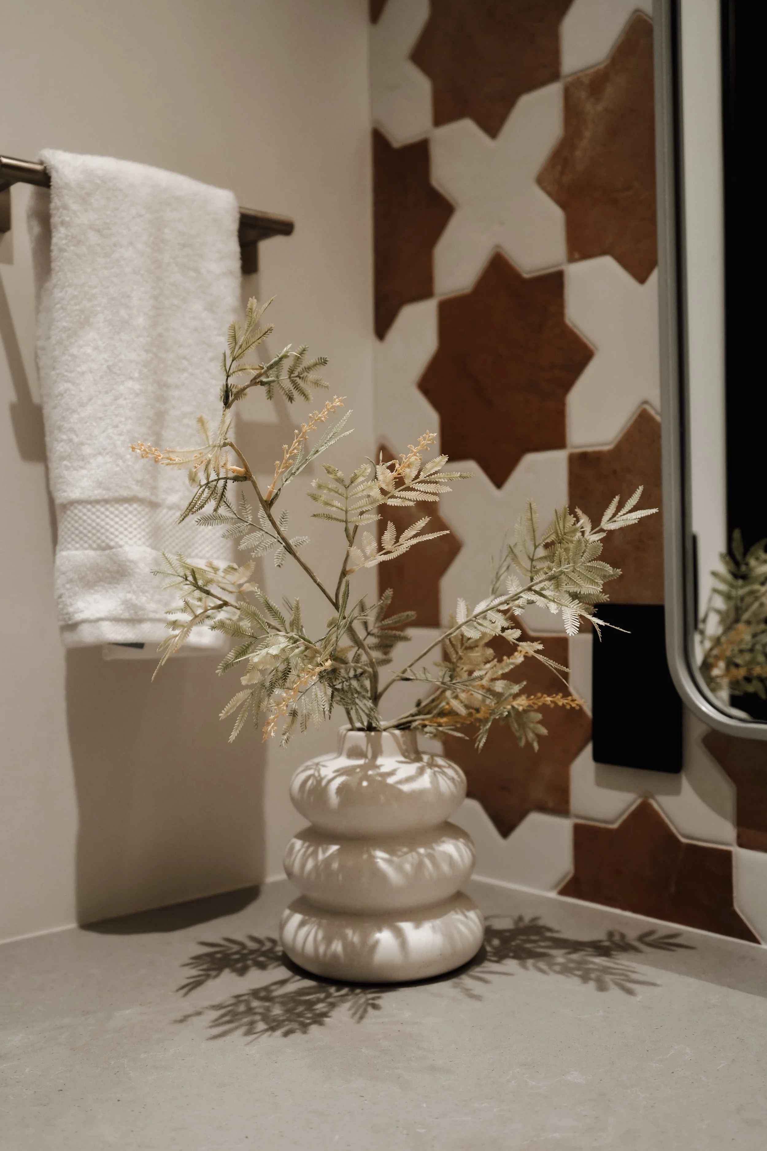 A white ceramic vase with three rounded sections containing a dried plant with light green leaves, placed on a bathroom countertop. Behind, a mirror reflects part of the wall decorated with star-shaped tiles. A white towel hangs on a metal towel bar 