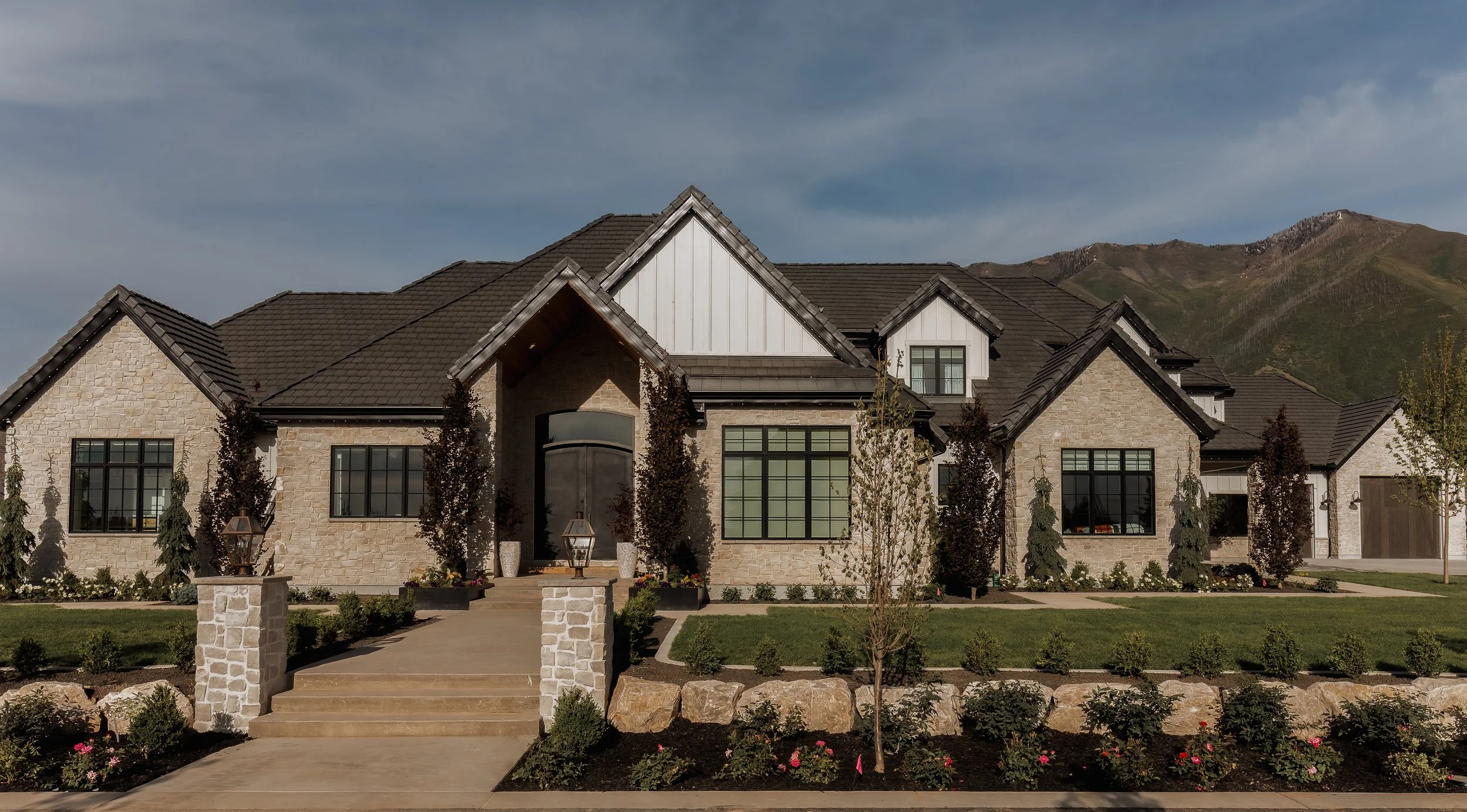A large modern house with stone and white siding, multiple gabled roofs, tall windows, and a landscaped front yard with shrubs, small trees, and a stone pathway.