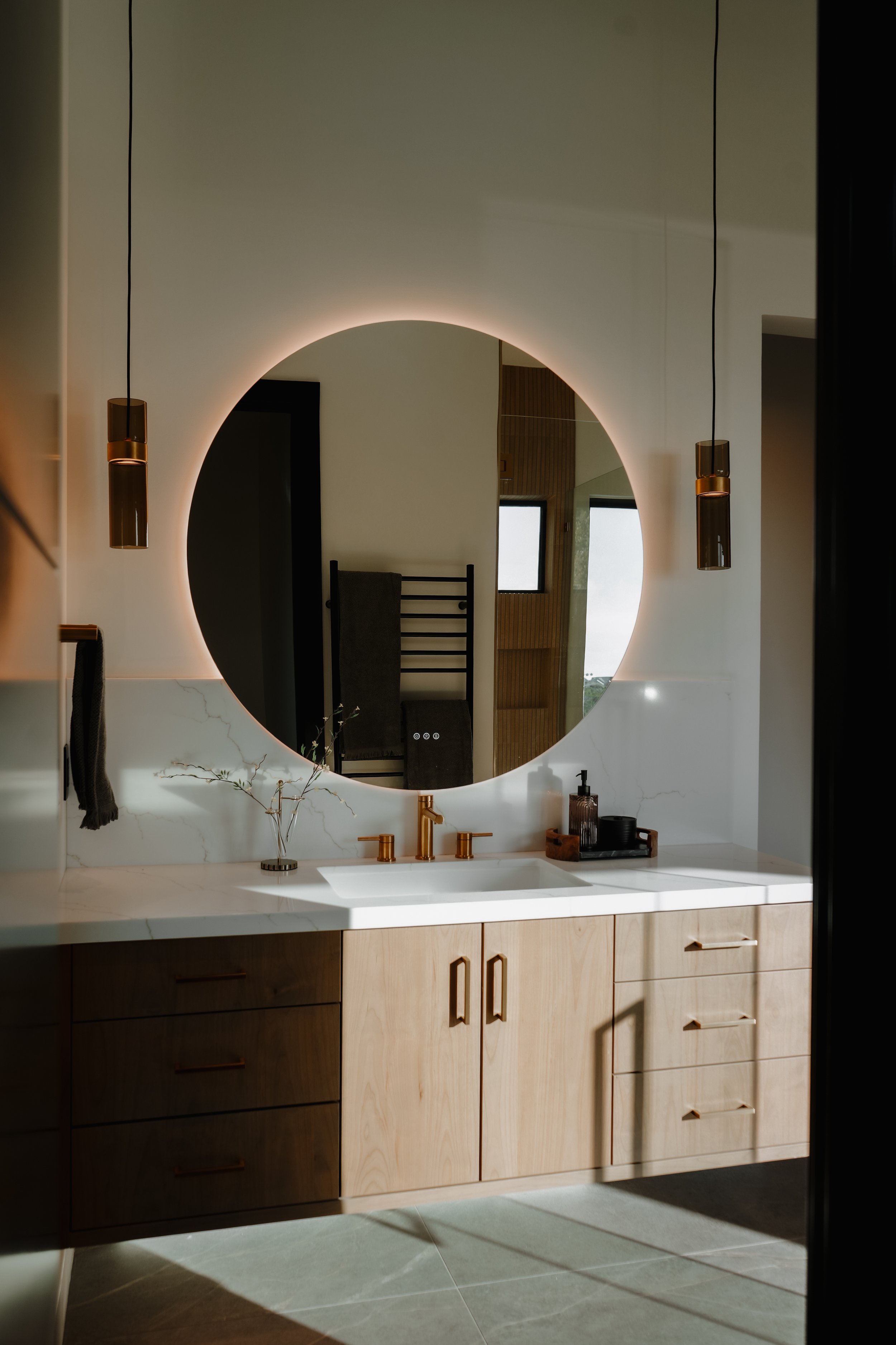 Modern bathroom vanity with a round mirror that has built-in lighting, a white marble countertop, and wooden cabinetry. Decor includes a small vase with branches, a soap dispenser, and toiletries tray. Two pendant lights hang on either side of the mi