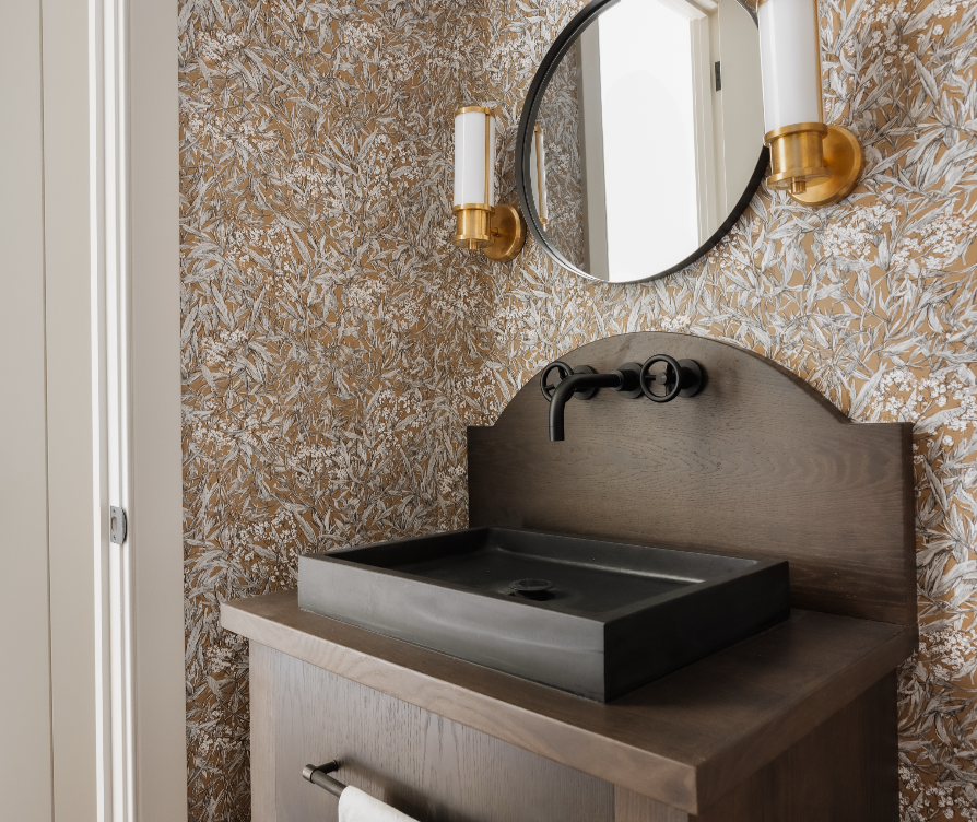 Modern bathroom vanity with a black rectangular vessel sink, gold wall sconces, a round mirror, and patterned wallpaper with leaf designs.