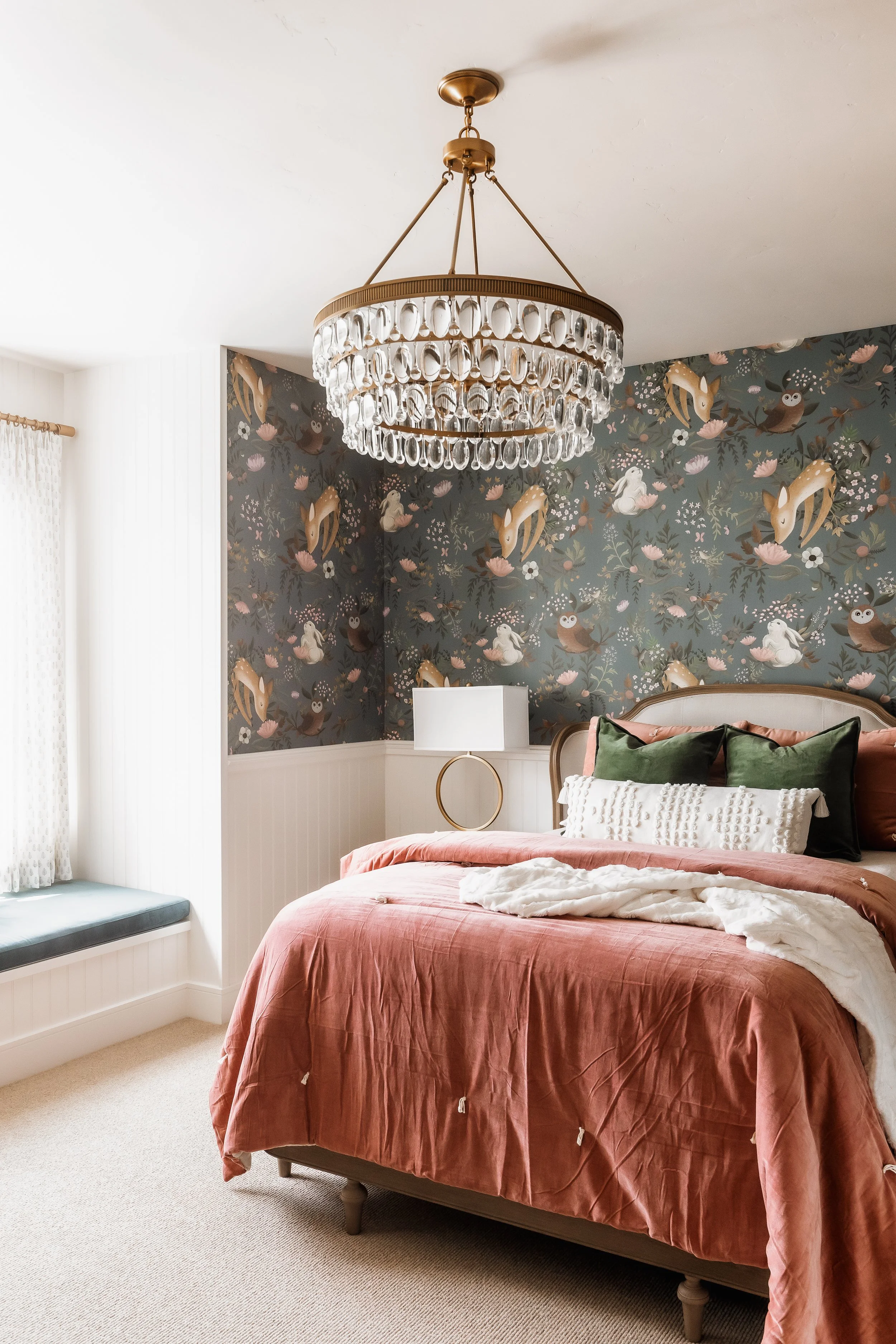 A bedroom with a bed, floral wallpaper, a chandelier, and a window with a window seat.