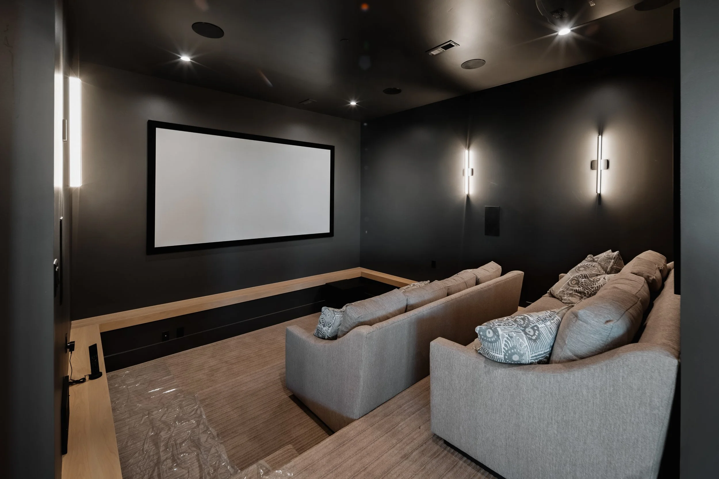 A private home theater room with two beige sofas facing a large blank screen, dim lighting, dark walls, and a carpeted floor.
