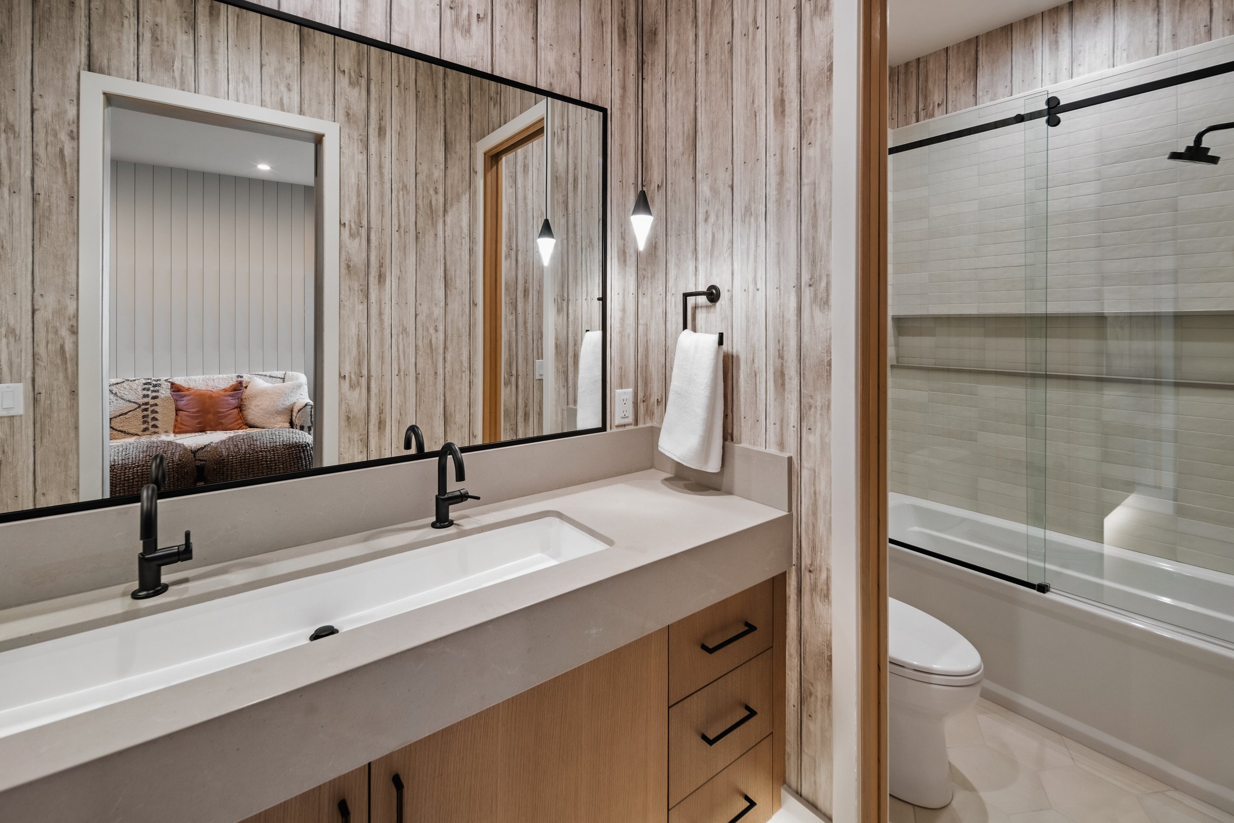Bathroom with wooden-paneled walls, large mirror, double sink vanity, wall-mounted towel rack, bathroom with bathtub and glass shower enclosure, toilet, and modern light fixtures.
