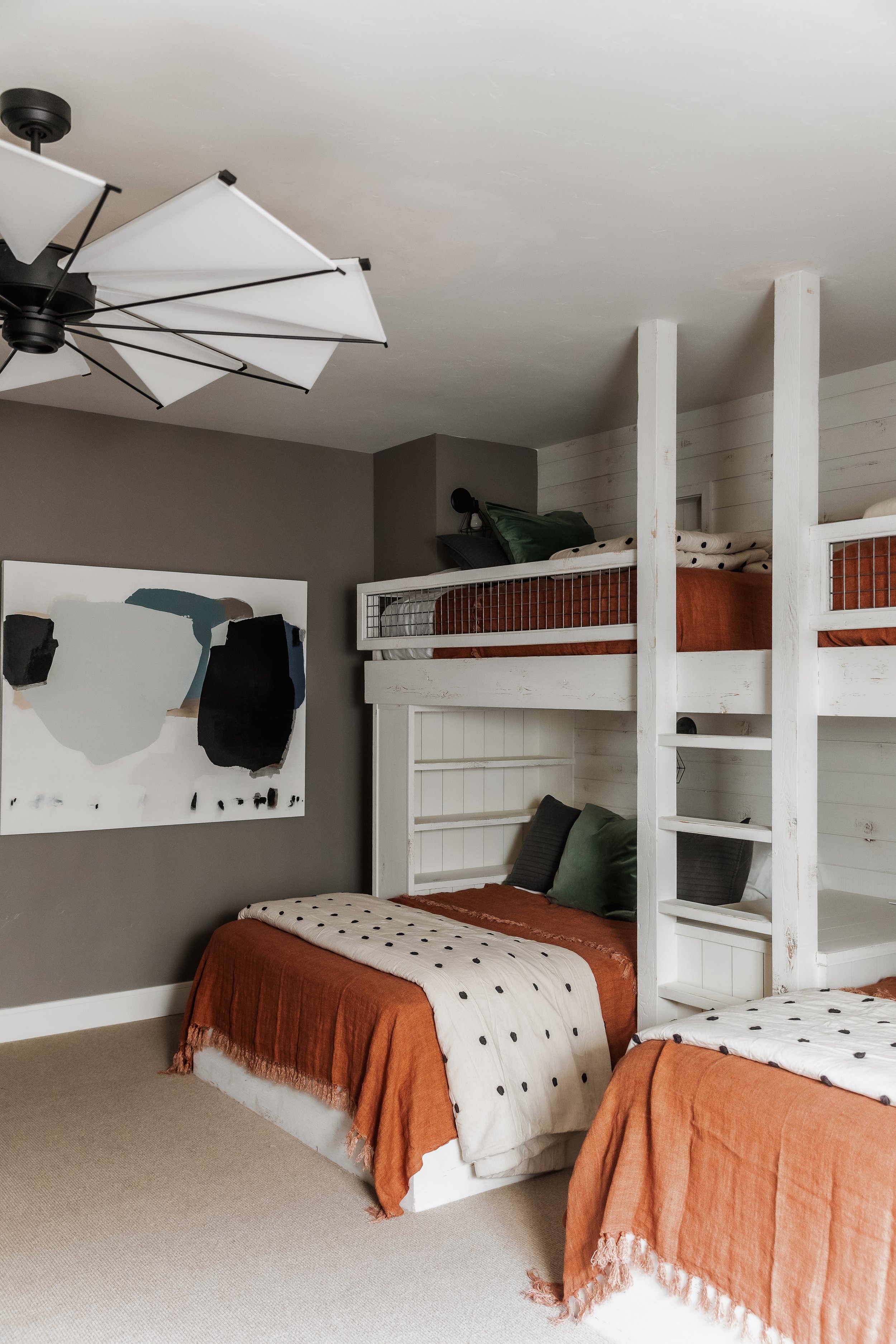 Room with two beds, one lower and one upper, with white wooden frame, green pillows, and rust-colored and white bedding with black polka dots, against a beige wall with a large abstract painting, ceiling fan, and a gray wall.