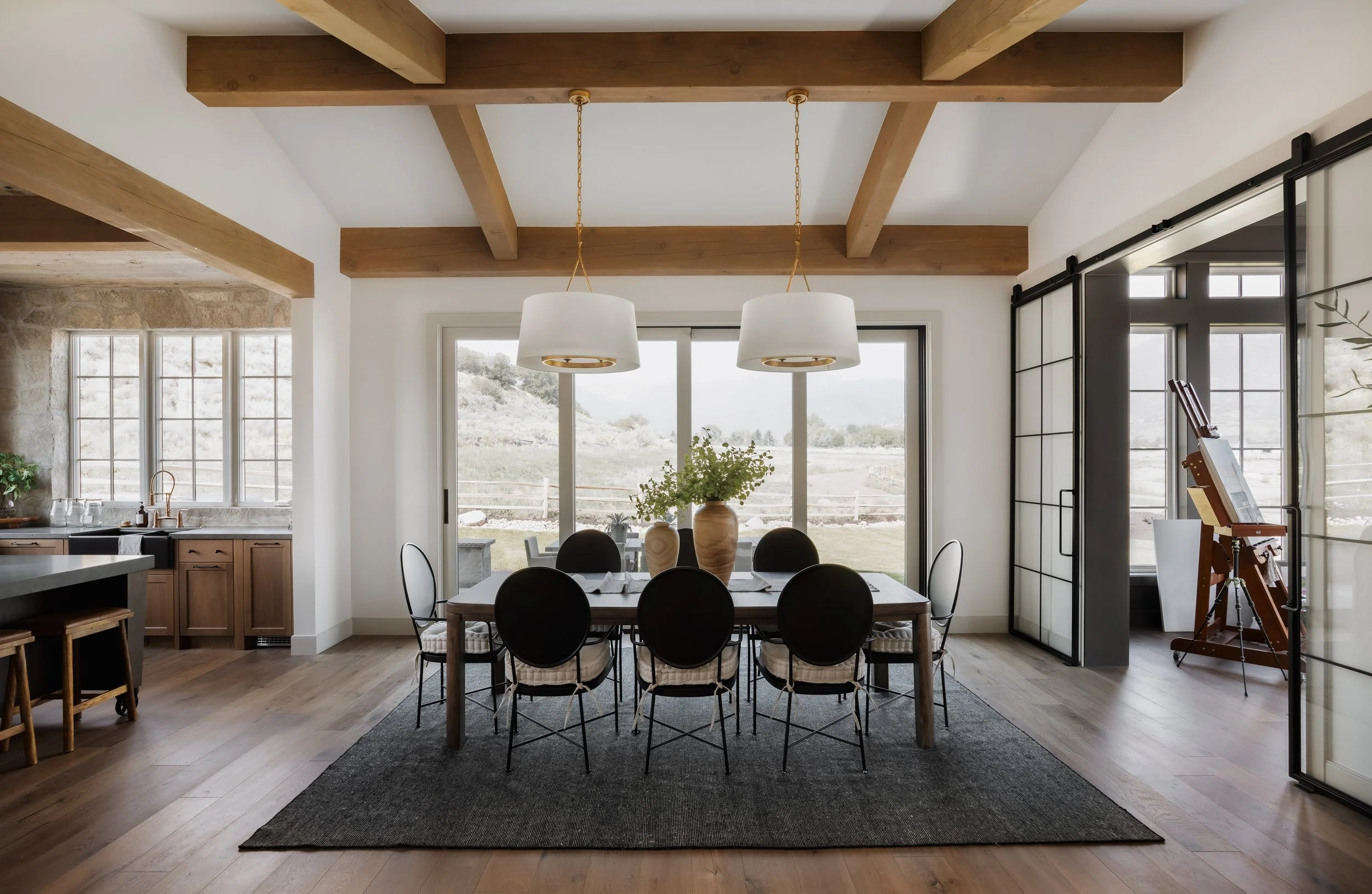 A bright dining room with a wooden table, six black chairs, and a large vase with greenery, illuminated by two hanging pendant lights, with a view of a backyard through sliding glass doors.