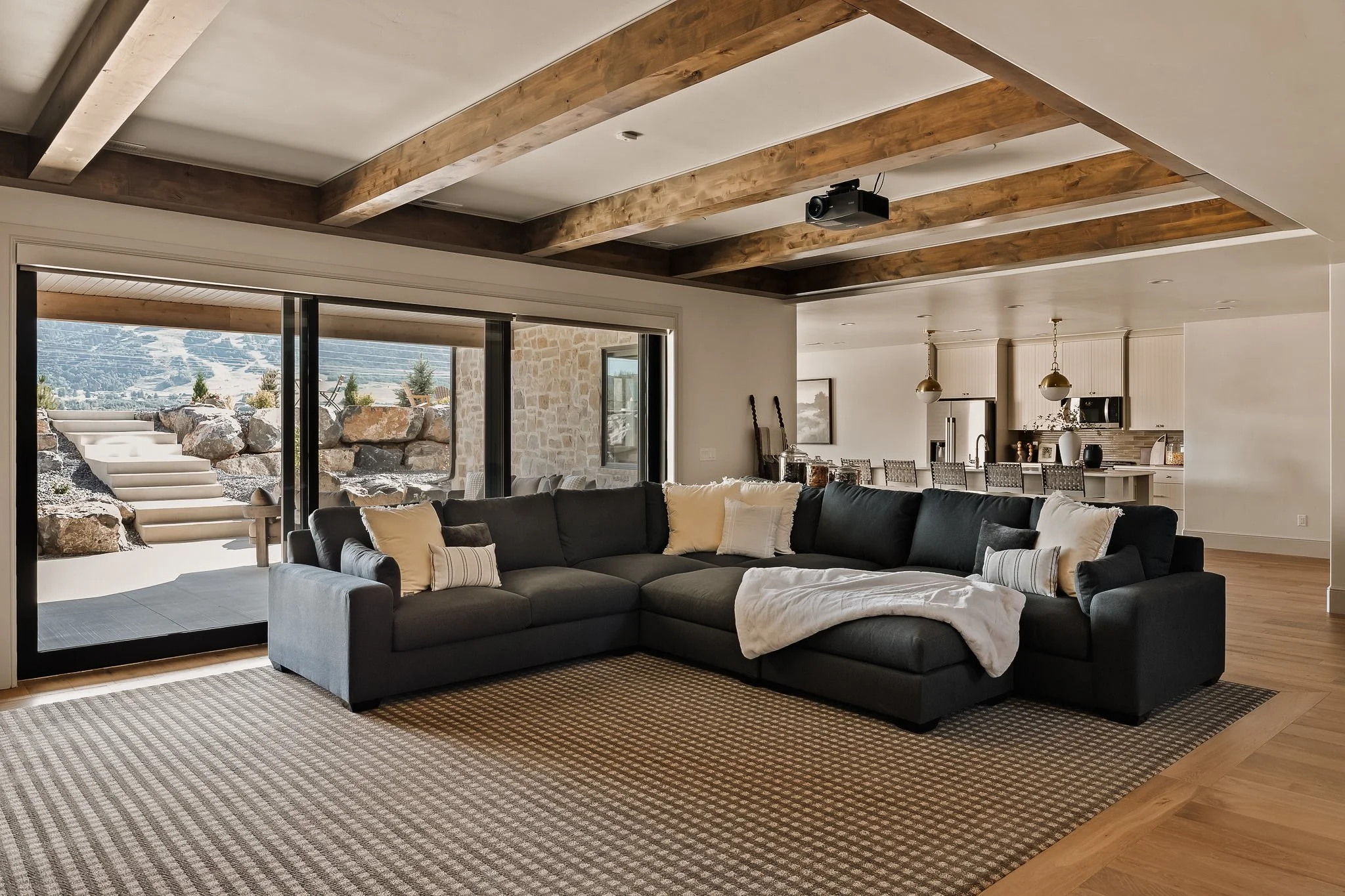 Living room with a large dark gray sectional sofa, pillows, and a blanket, overlooking a stone patio with stairs and a scenic outdoor view through sliding glass doors, in a modern open-concept home with exposed wooden ceiling beams.