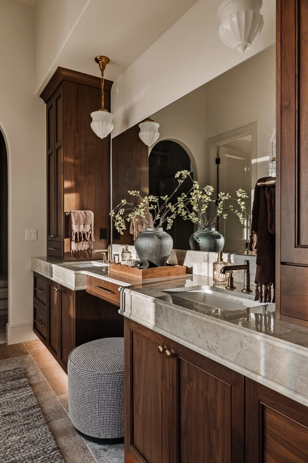 Bathroom vanity with wood cabinets, marble countertop, large mirror, decorative vases with branches, and wall-mounted light fixtures.