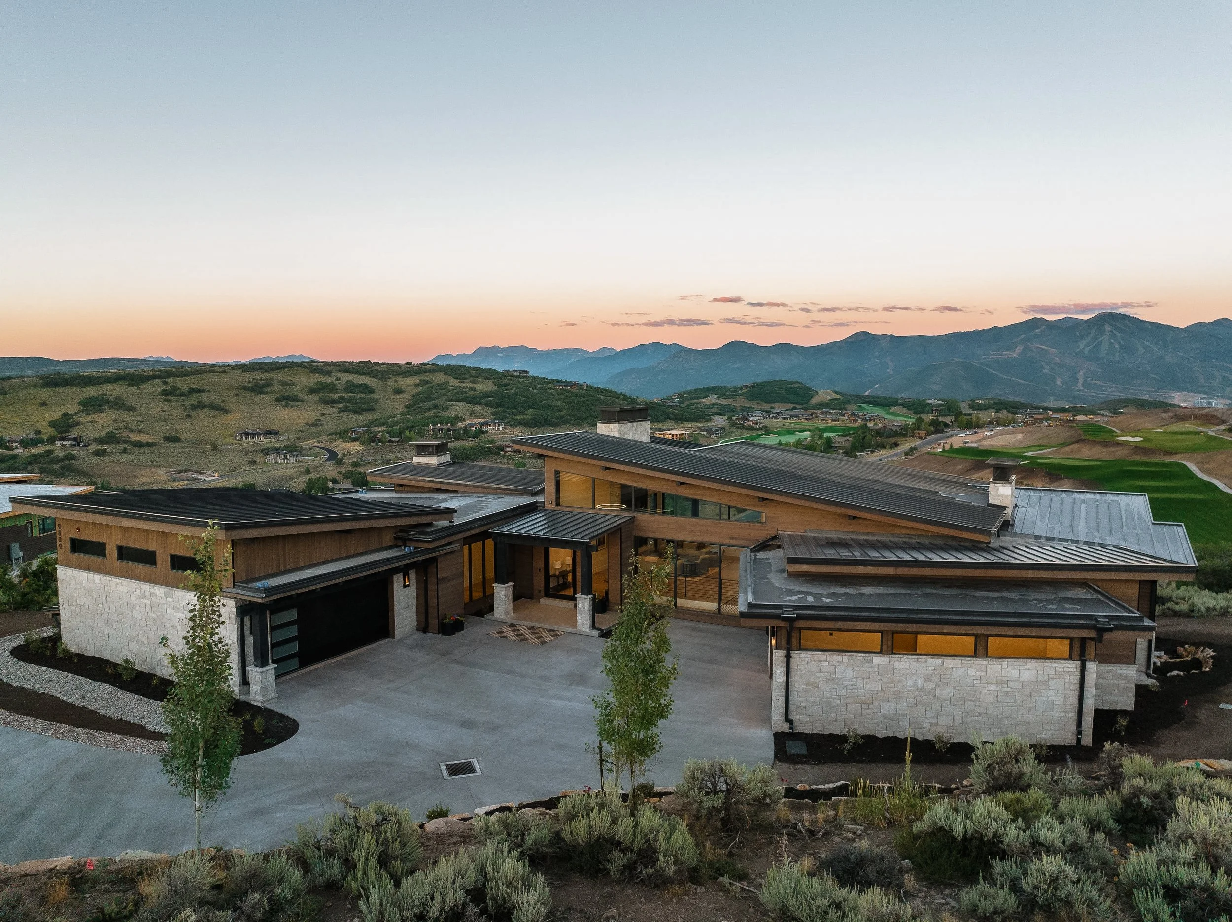 Modern house with metal roof and stone accents in a hilly landscape at dusk, mountains in the background.