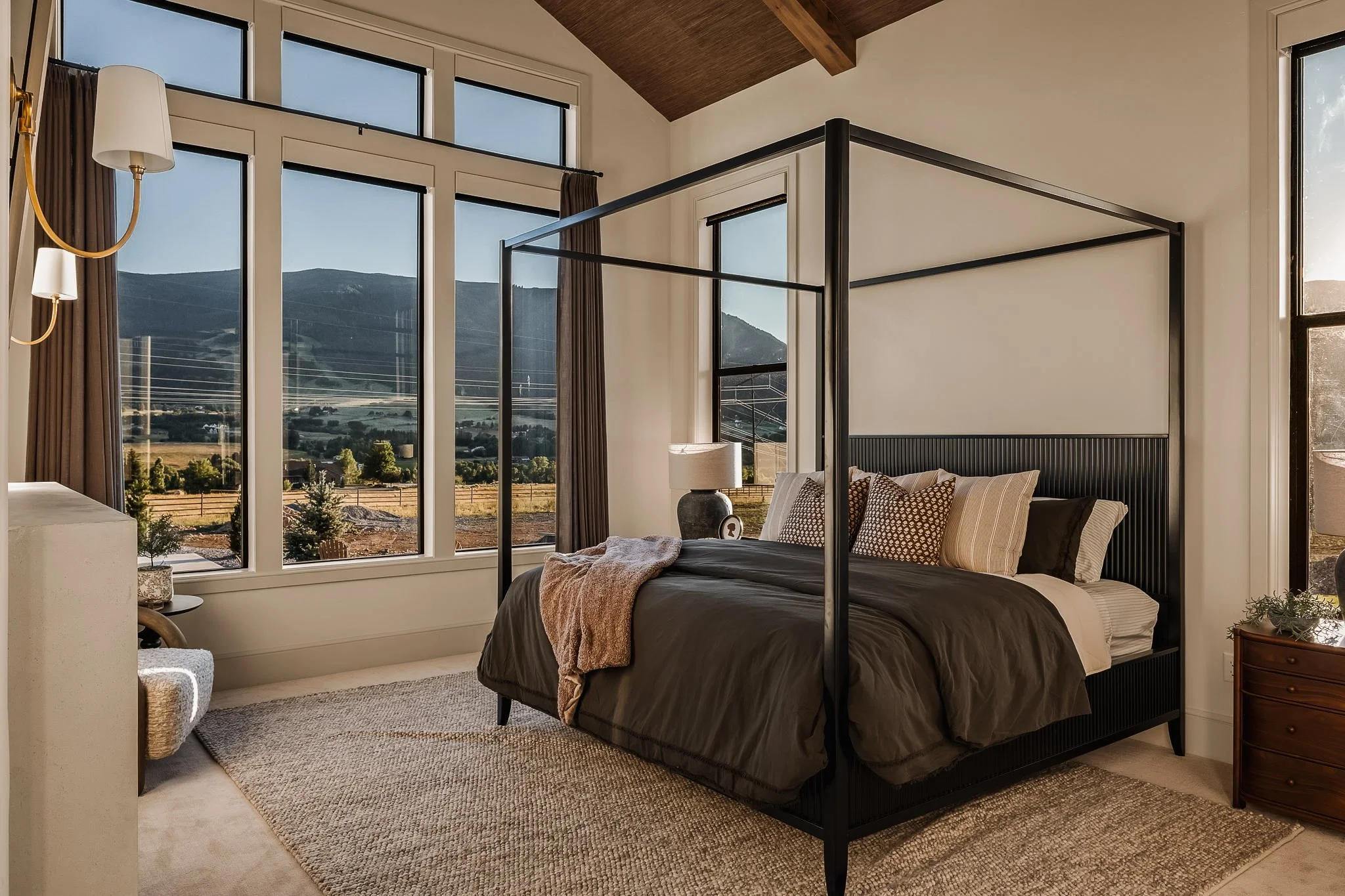 A bedroom with large windows showing mountains outside, a black metal four-poster bed with beige and brown pillows, a bedside table with a lamp, and a beige rug on the floor.