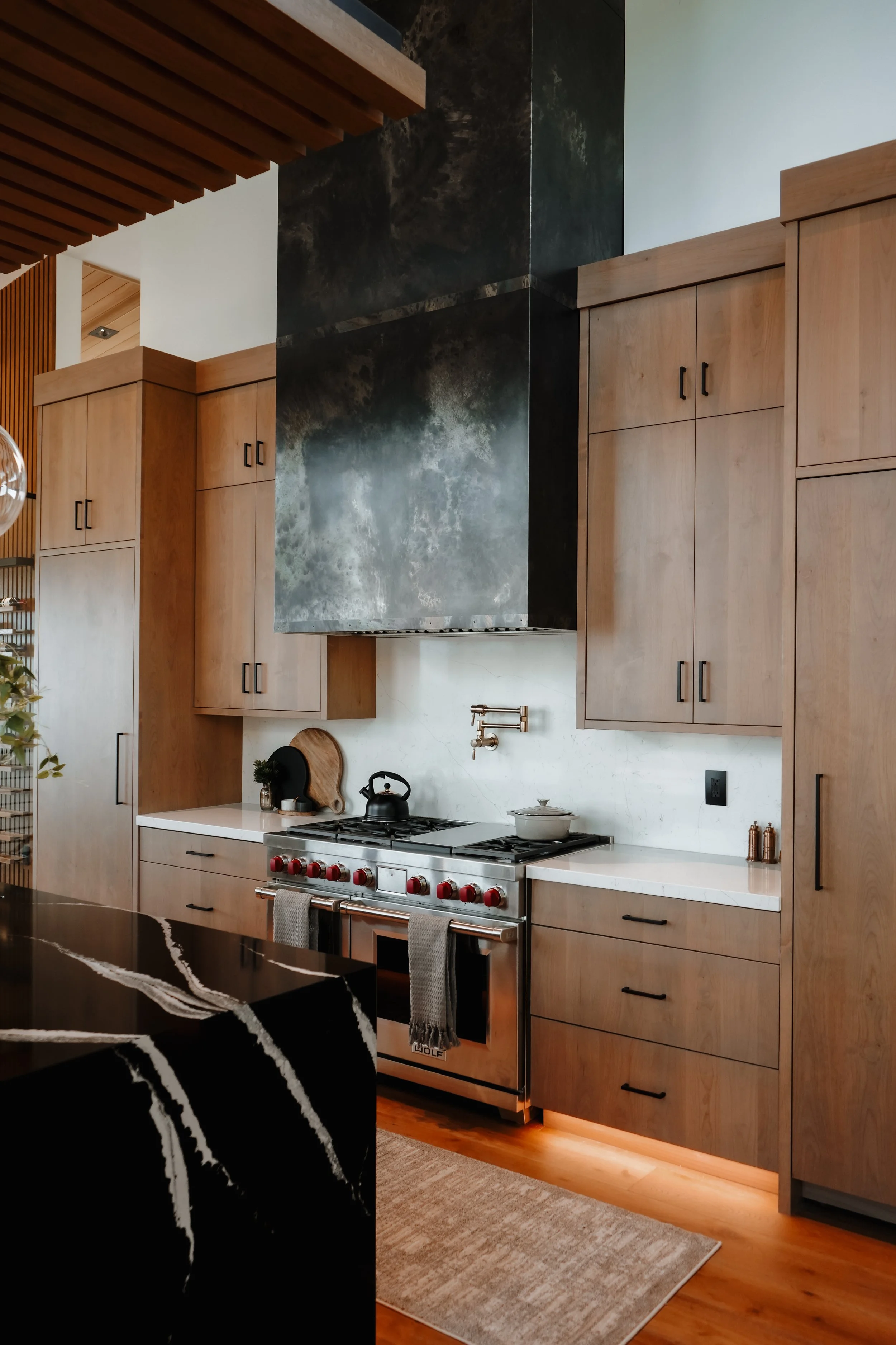 A modern kitchen with light wooden cabinets, a stainless steel stove with red knobs, a black kettle, and cutting boards. There is a marble backsplash and a vent hood above the stove. The kitchen features a combination of natural wood tones and black 