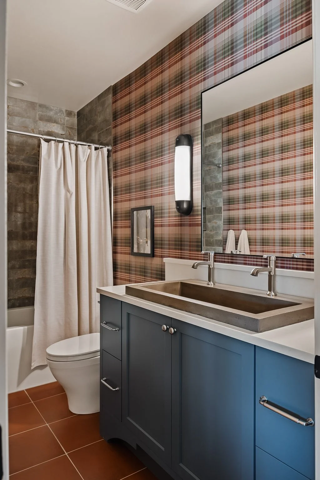 A bathroom with plaid wallpaper, a large mirror, a double sink vanity with blue cabinetry, a rectangular countertop sink, a wall-mounted light fixture, a framed picture, a toilet, and a shower with a beige curtain.