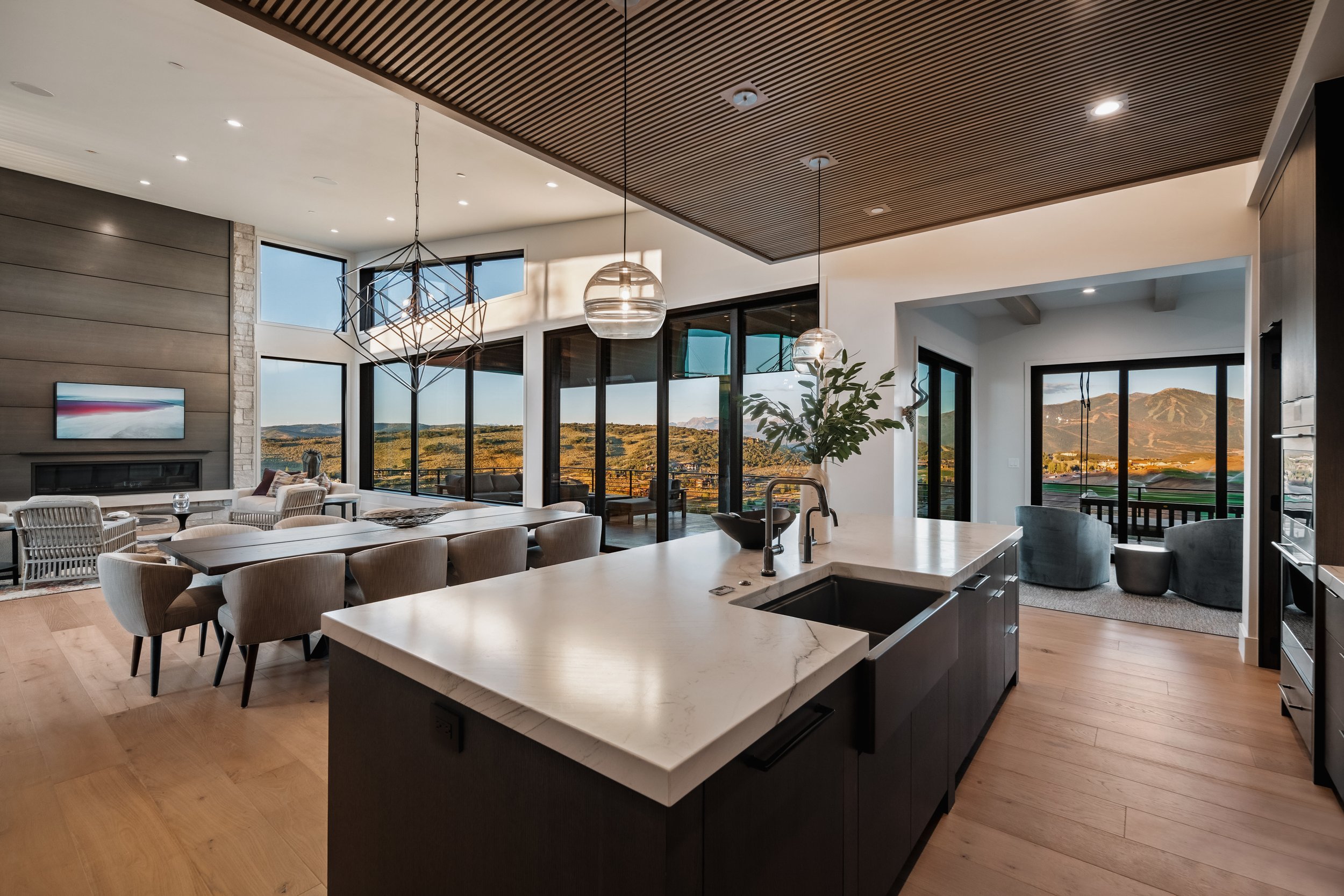 Open-concept living and dining area with large windows offering a mountain view, contemporary furniture, a kitchen island with a sink, hanging pendant lights, and a cozy nook with armchairs and a view of the outdoors.