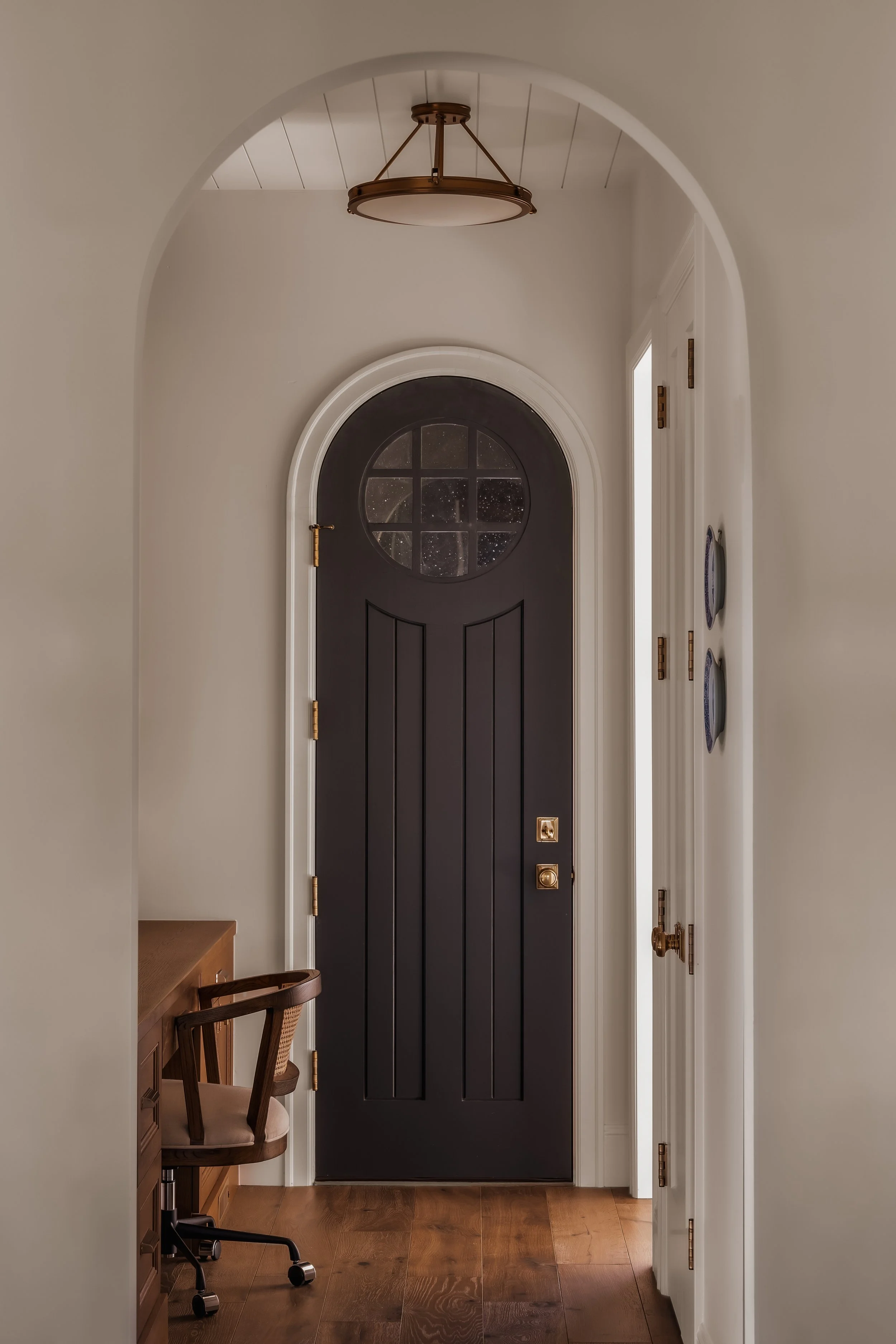View of a home's front door with black paint and a semi-circular window, surrounded by white trim. The interior features white walls, wooden flooring, and a ceiling light fixture. To the left, there's a wooden desk with a matching chair, and to the r