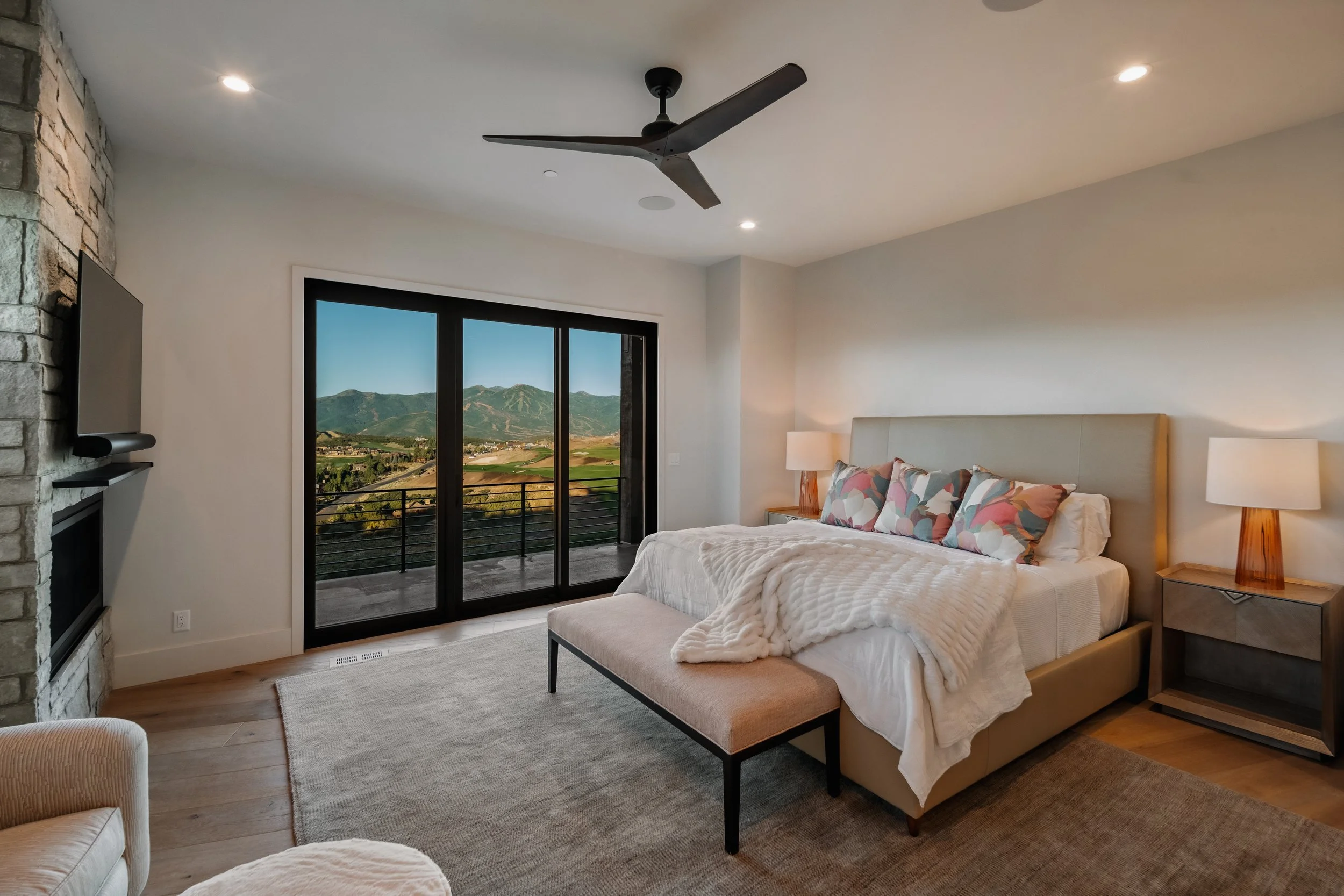 Modern bedroom with sliding glass door leading to a balcony, mountain view, beige bed with colorful pillows, white throw blanket, two wooden nightstands with lamps, ceiling fan, stone fireplace, and a TV.