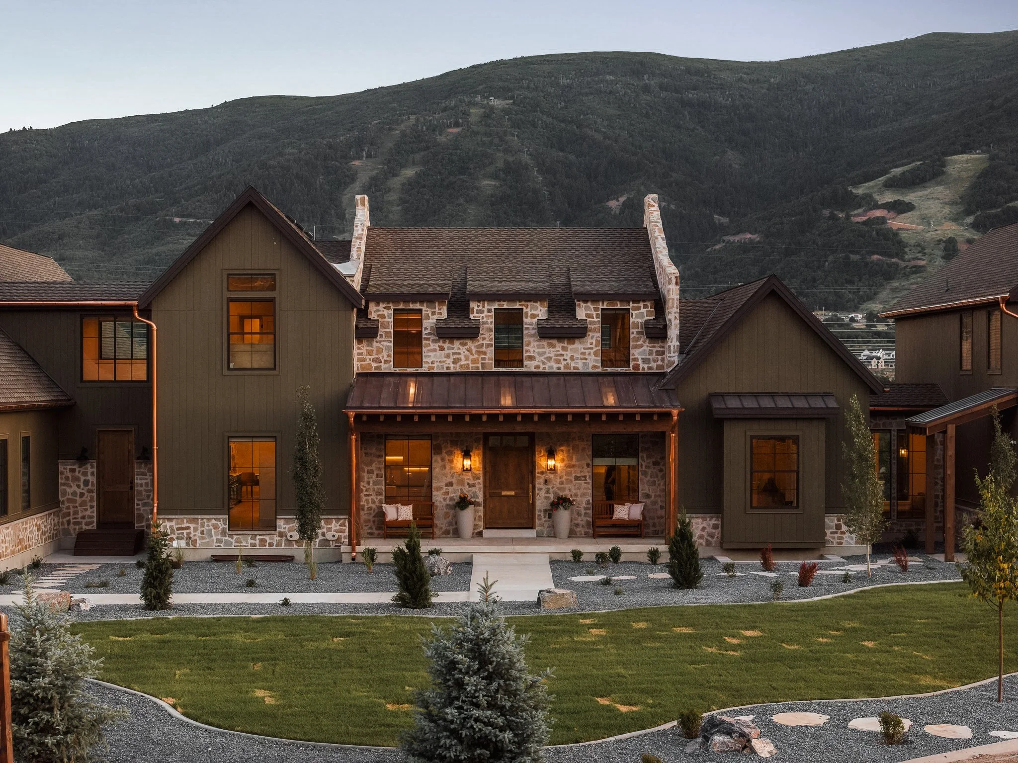 A modern house with a stone and wood exterior, situated in a mountainous area, illuminated with warm lights at dusk.