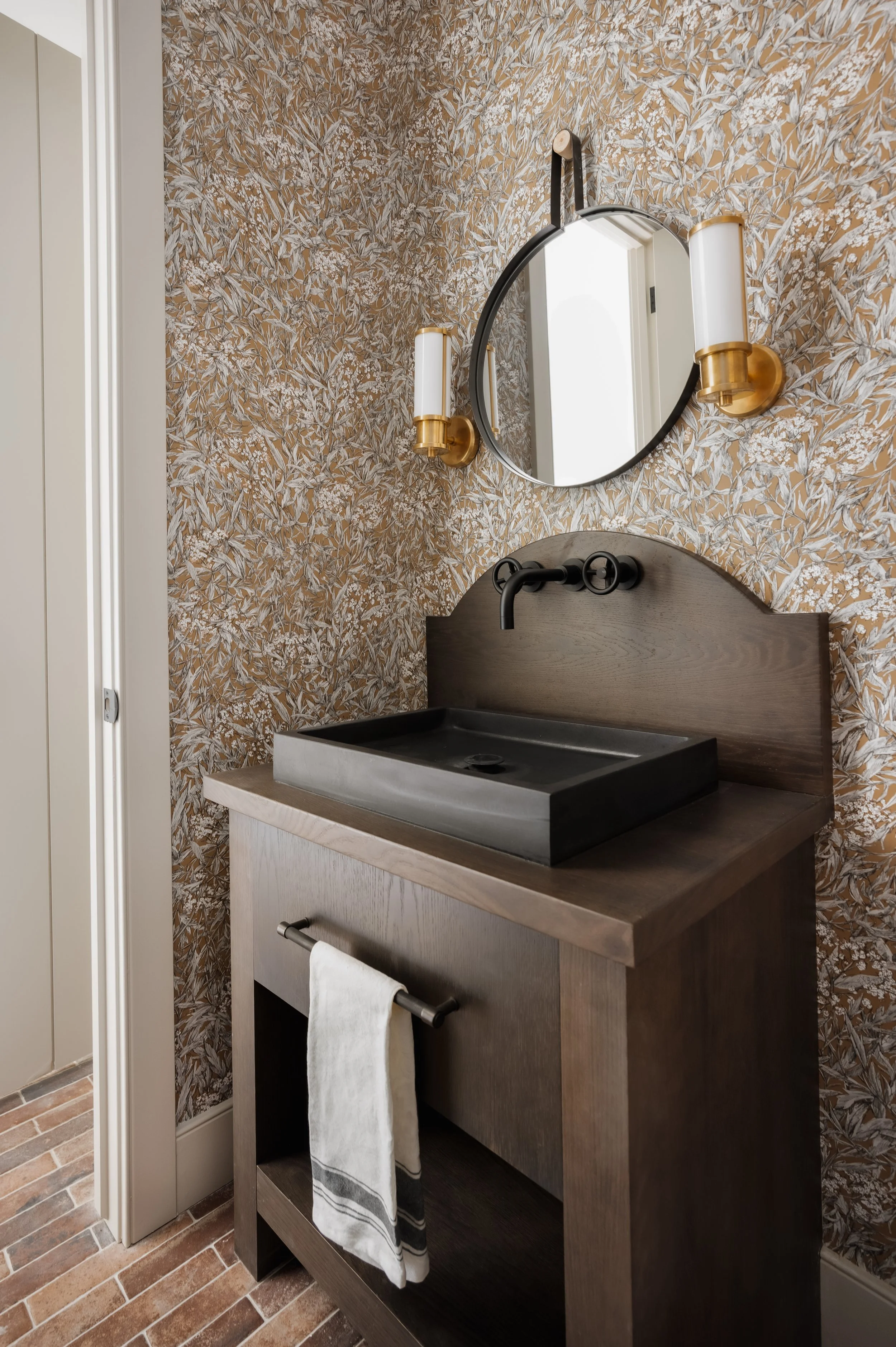 A bathroom vanity with a black rectangular vessel sink, a wooden cabinet, a round mirror above, gold and white wall-mounted light fixtures, and a floor with brick-patterned tiles.