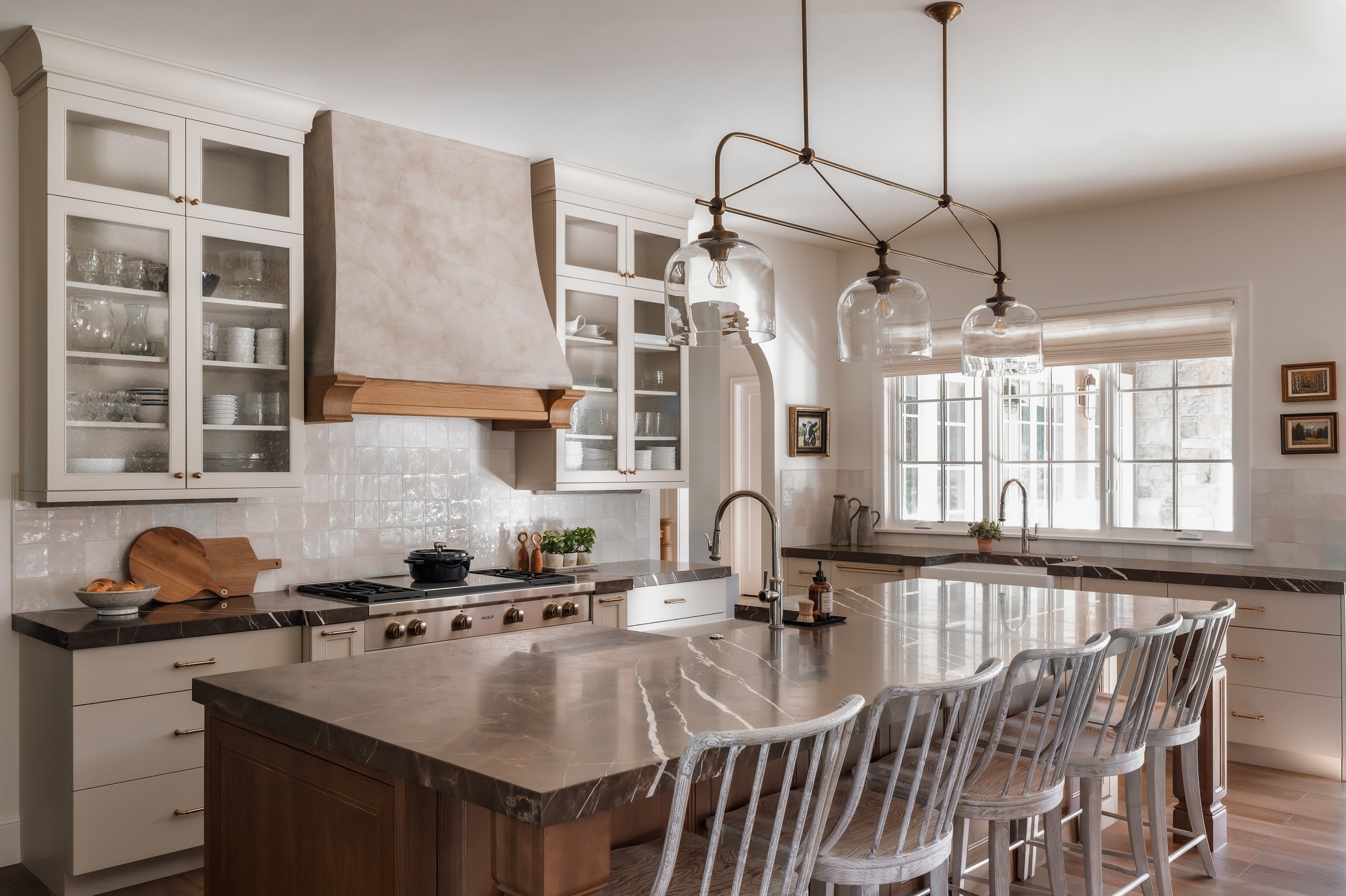 A spacious kitchen with white cabinets, a large marble island, and natural light from multiple windows, featuring modern lighting fixtures hanging above the island.
