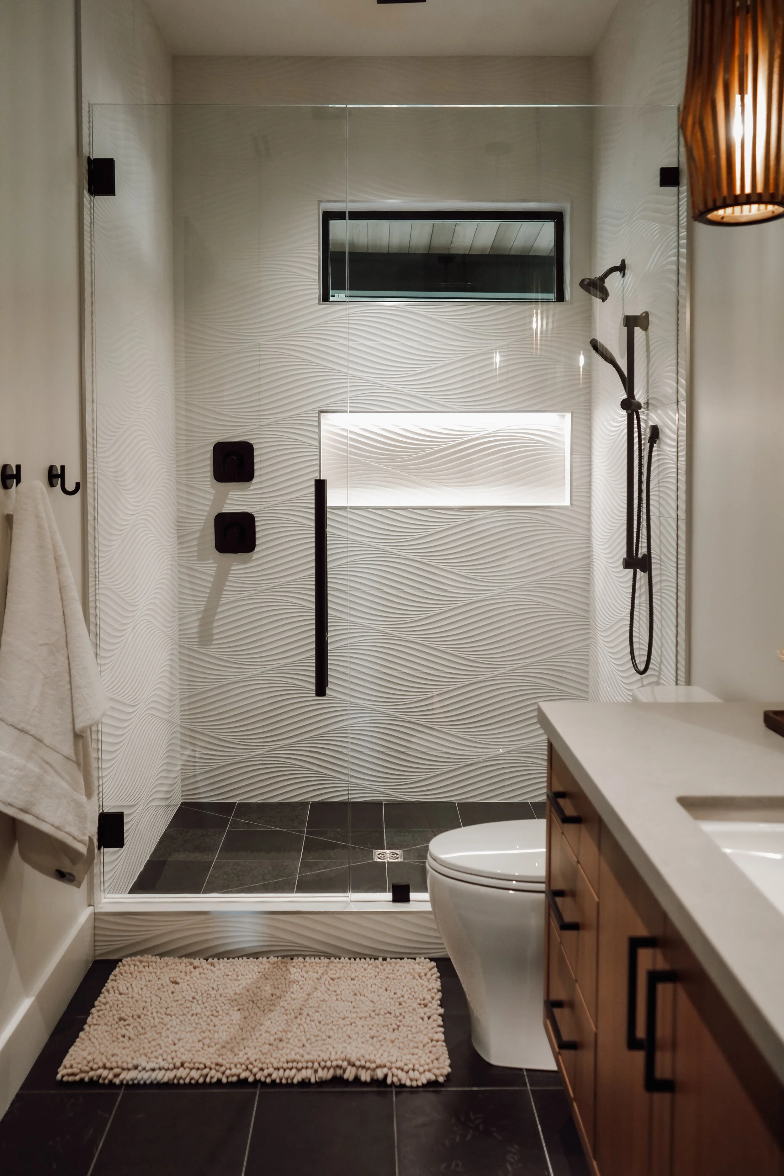 Modern bathroom with a glass-enclosed walk-in shower featuring textured white wall tiles, a small window above, and black fixtures. A beige bath mat is on dark tiled floor, with a white toilet and wooden vanity with a white countertop partially visib