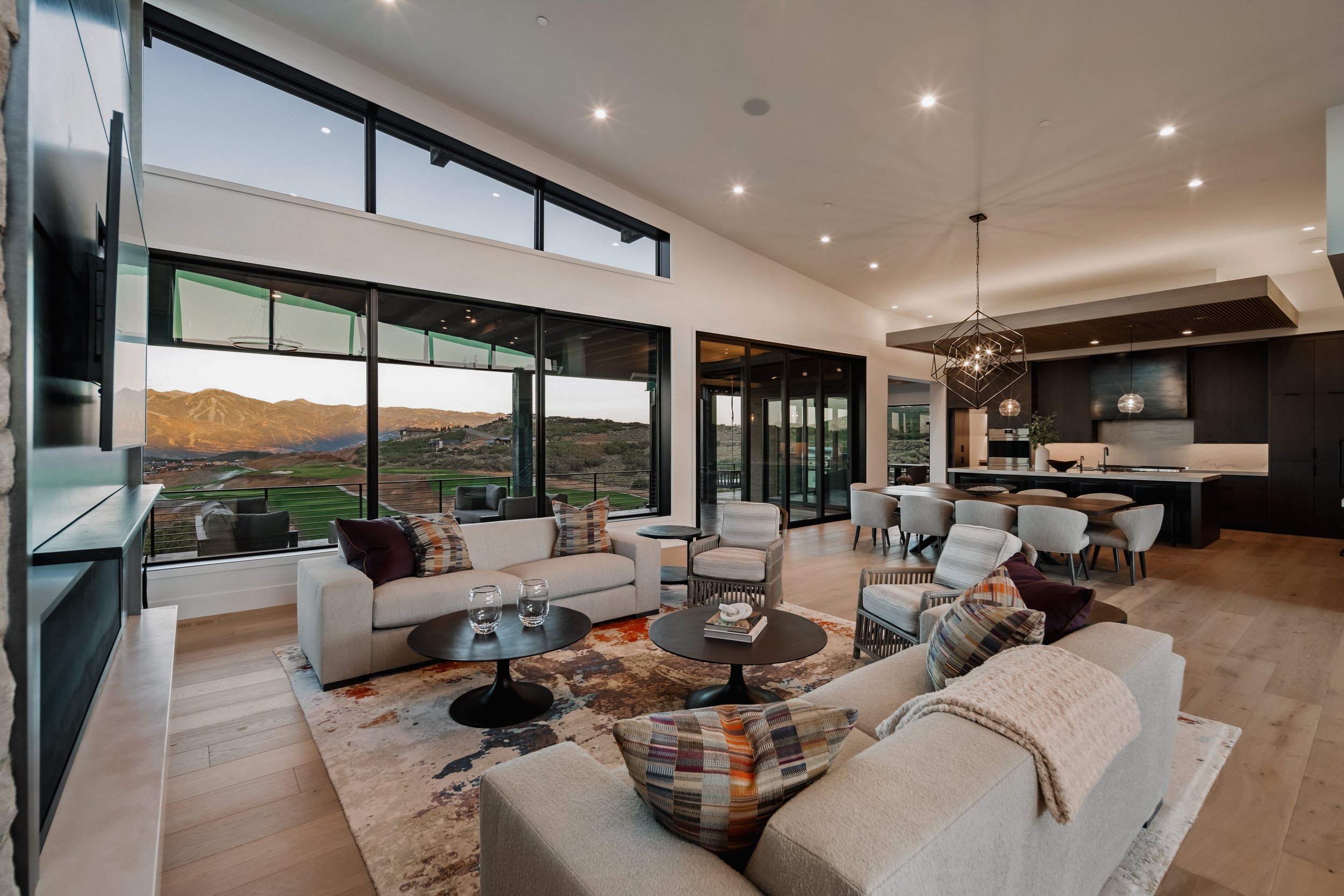 Interior view of a modern, open-concept living room and kitchen with large windows showcasing mountain and landscape views, furnished with sofas, armchairs, coffee tables, a dining table, and contemporary lighting fixtures.