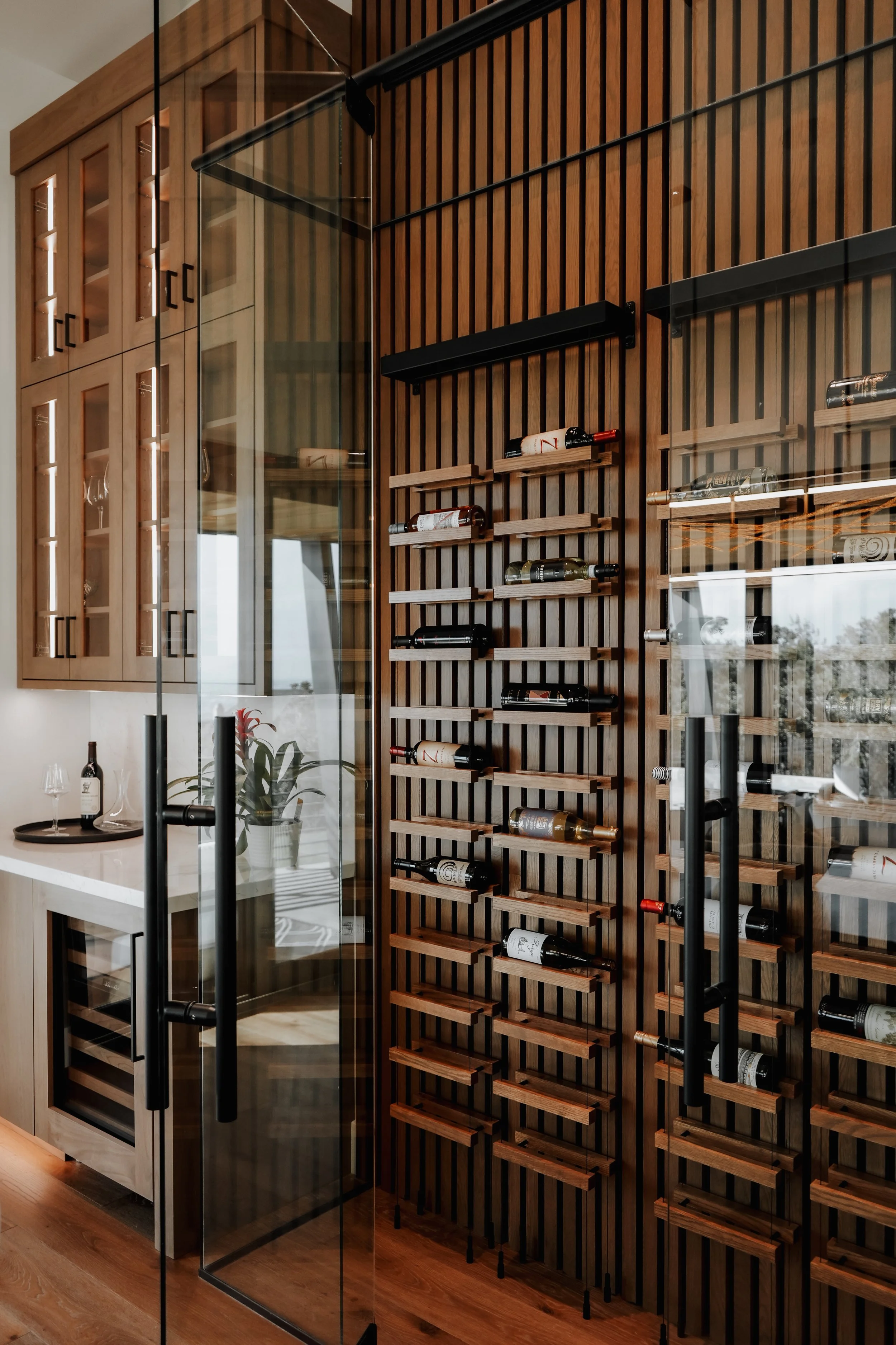 Interior view of a wine cellar with glass door, wooden wine racks holding bottles, and a built-in cabinet with glass doors displaying glasses.