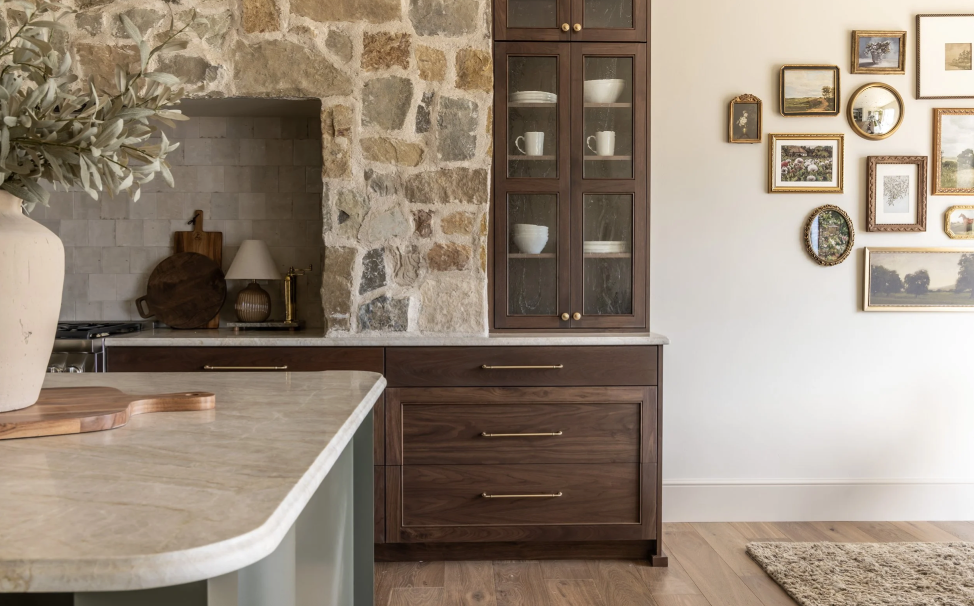 A kitchen with a stone wall and a wooden cabinet with glass doors, displaying white dishes. A framed gallery wall with various pictures is on the right, and a marble countertop with a vase and cutting board is in the foreground.