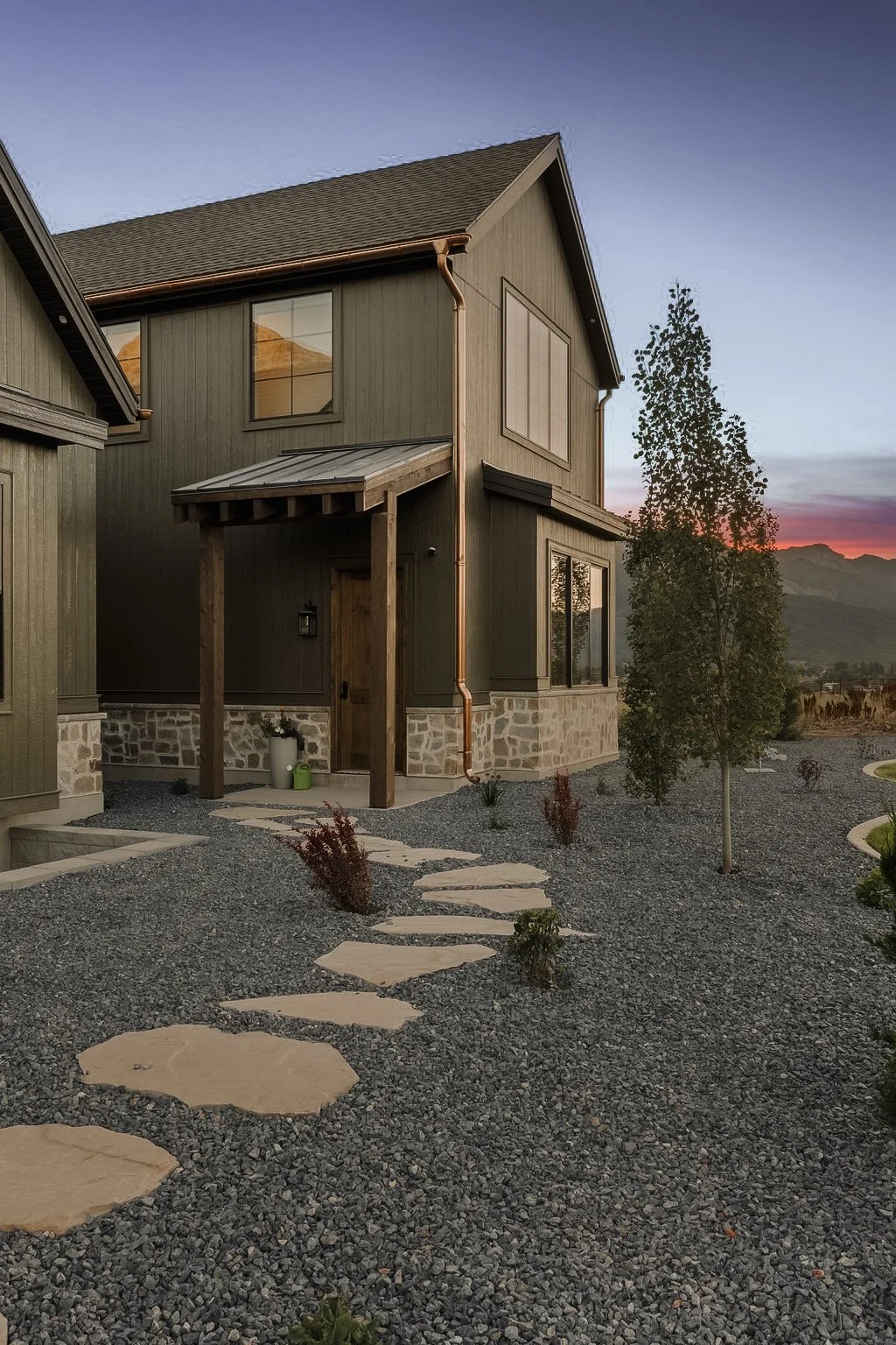 A modern two-story house with dark gray siding, stone foundation, and large windows. The front yard features a stone pathway with pea gravel and small plants, with a tree on the right and mountain scenery in the background during sunset.