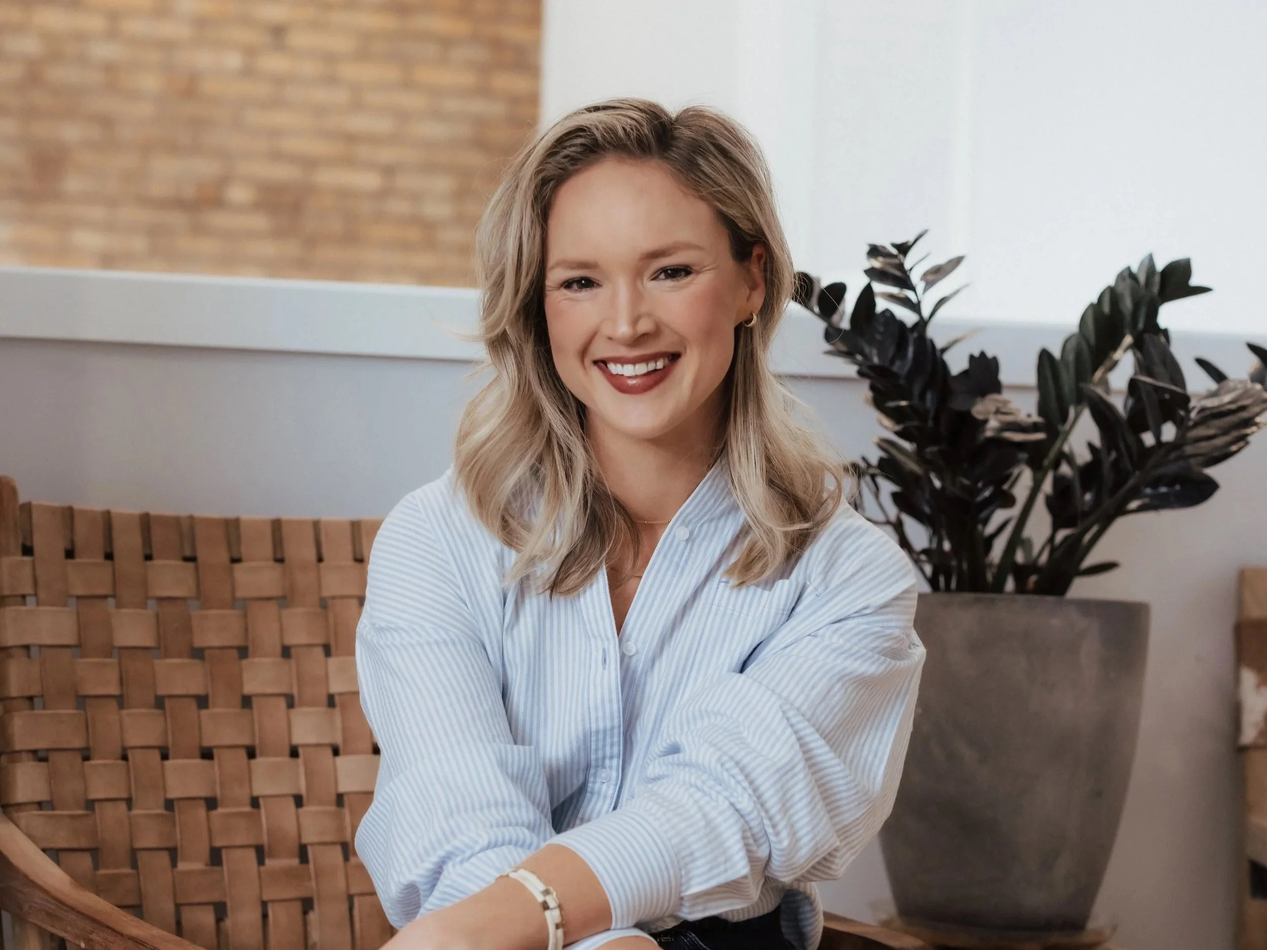 A young woman with blonde hair styled in loose waves, wearing a light blue button-up shirt, smiling and sitting on a wicker chair indoors next to a large potted plant with dark green leaves.