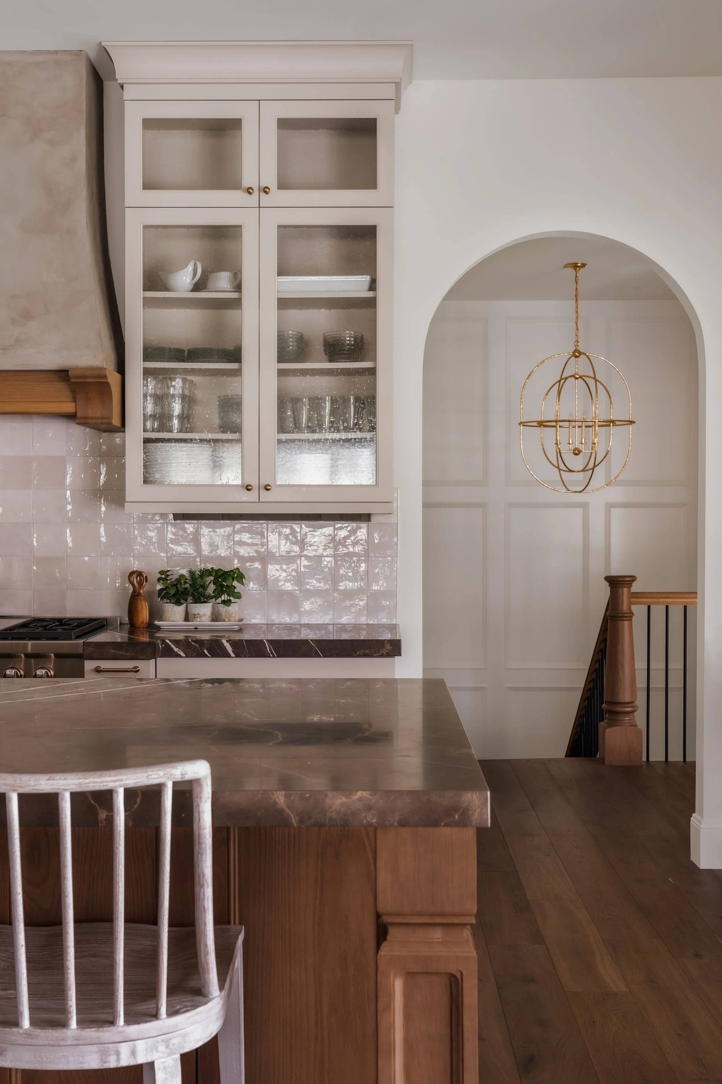 Kitchen with beige tiled backsplash, glass-front cabinet, marble countertop, wooden island, and a modern gold hanging chandelier.