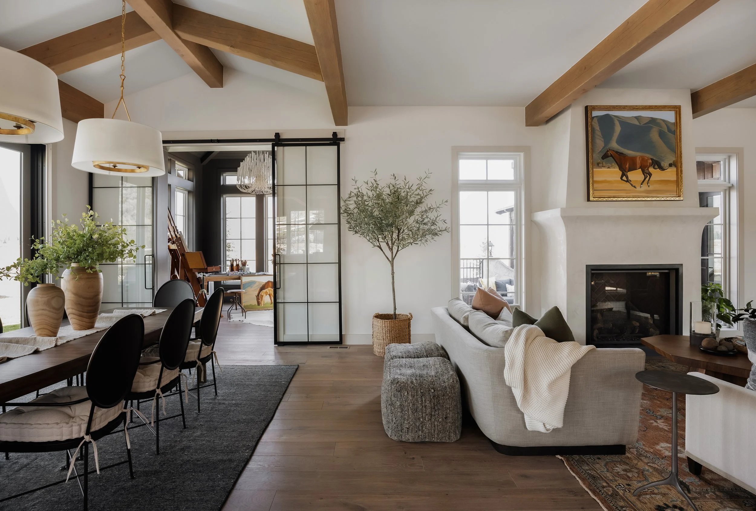Interior of a modern living room with a dining area and fireplace, featuring wooden beams on the ceiling, a large window, and minimalistic decor.