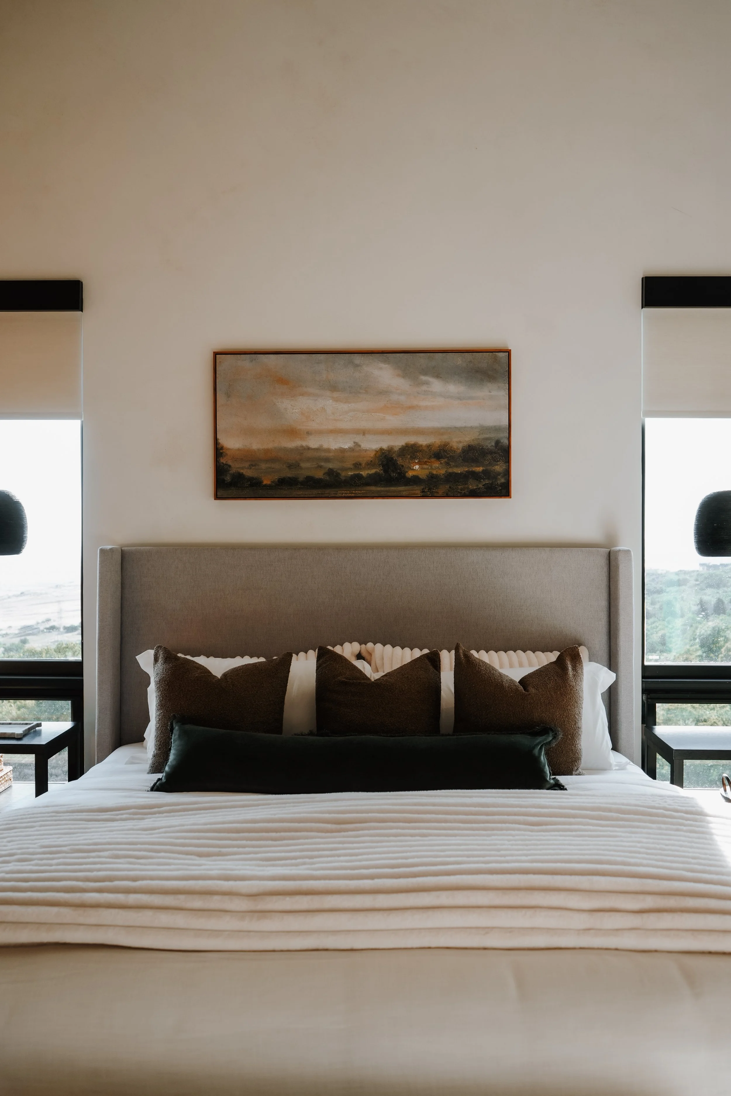 A neatly made bed with beige headboard, layered pillows, and a white textured blanket in a modern bedroom with two windows and a landscape painting above the bed.