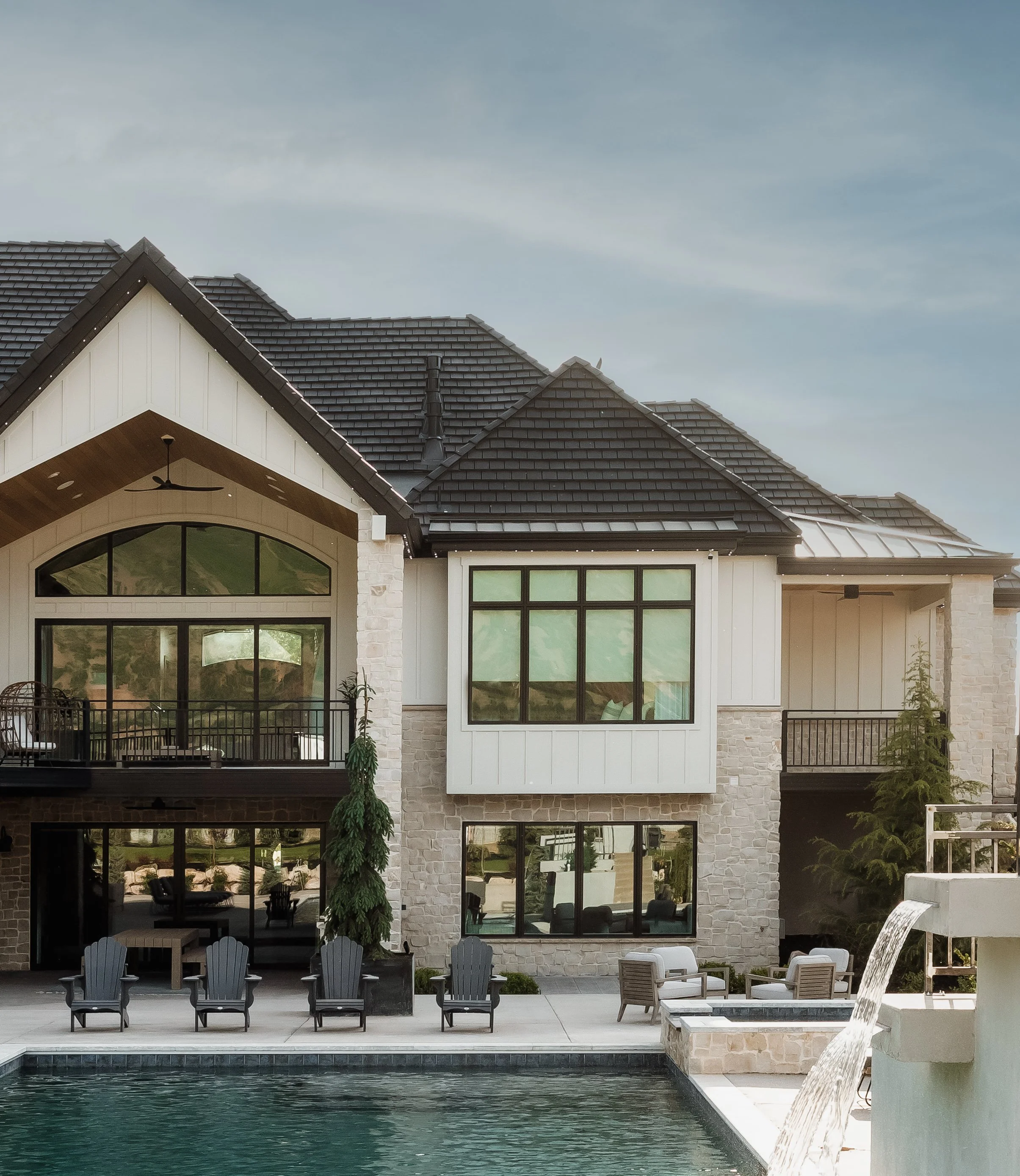 Modern multi-story house with large windows, a pool in the foreground, outdoor seating, and a small waterfall feature, under a partly cloudy sky.