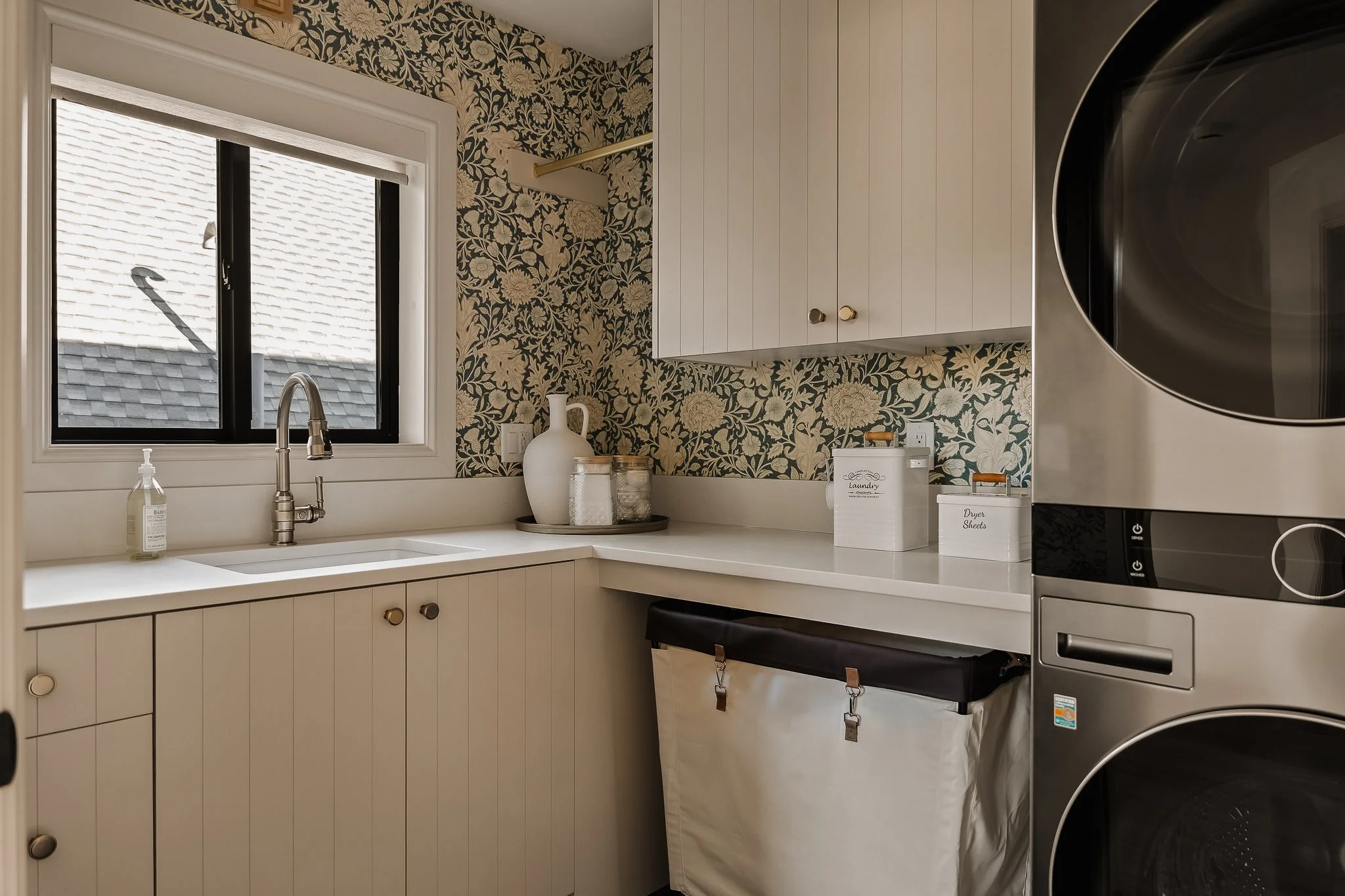 Laundry room with a window, floral wallpaper, white cabinets, a laundry basket, and a washing machine.