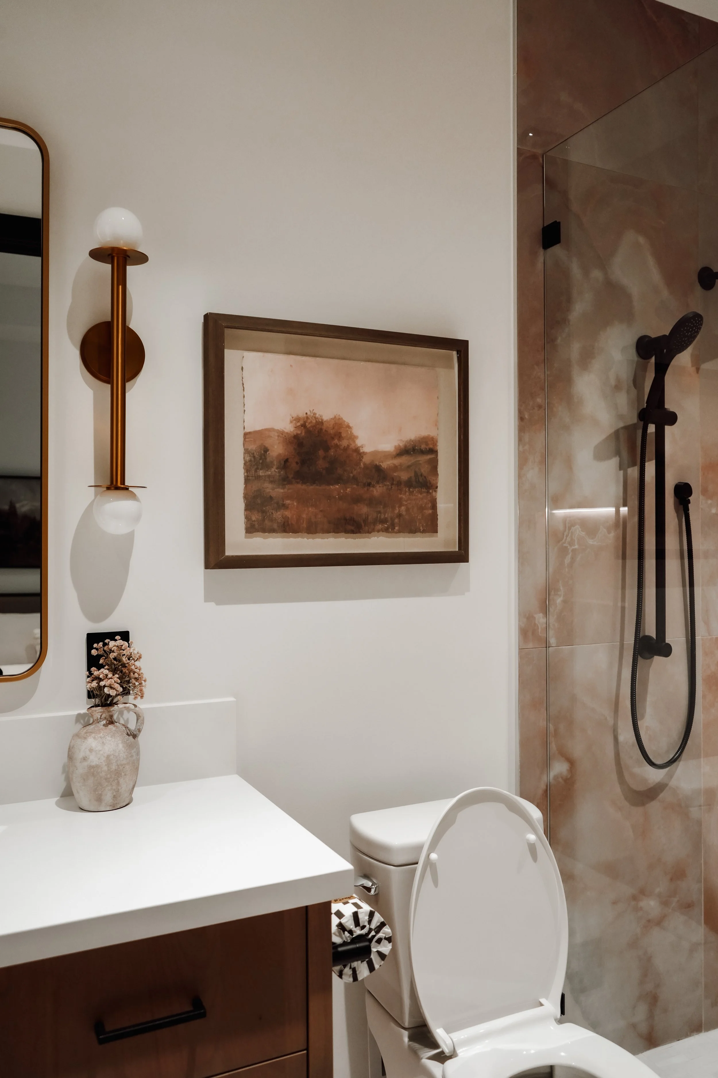A modern bathroom with a white toilet, a wooden vanity with a white countertop, a potted flower, a framed landscape painting, a mirror, a gold and white wall light, and a shower area with beige and brown marble tiles and a black showerhead.