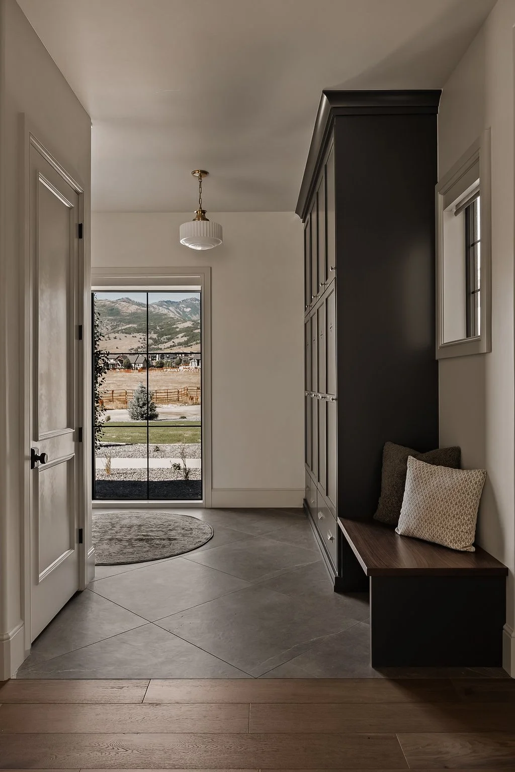 Entryway with a bench and pillows on the right, a large window showing mountains outside, a round rug on tile floor, and a ceiling light fixture.