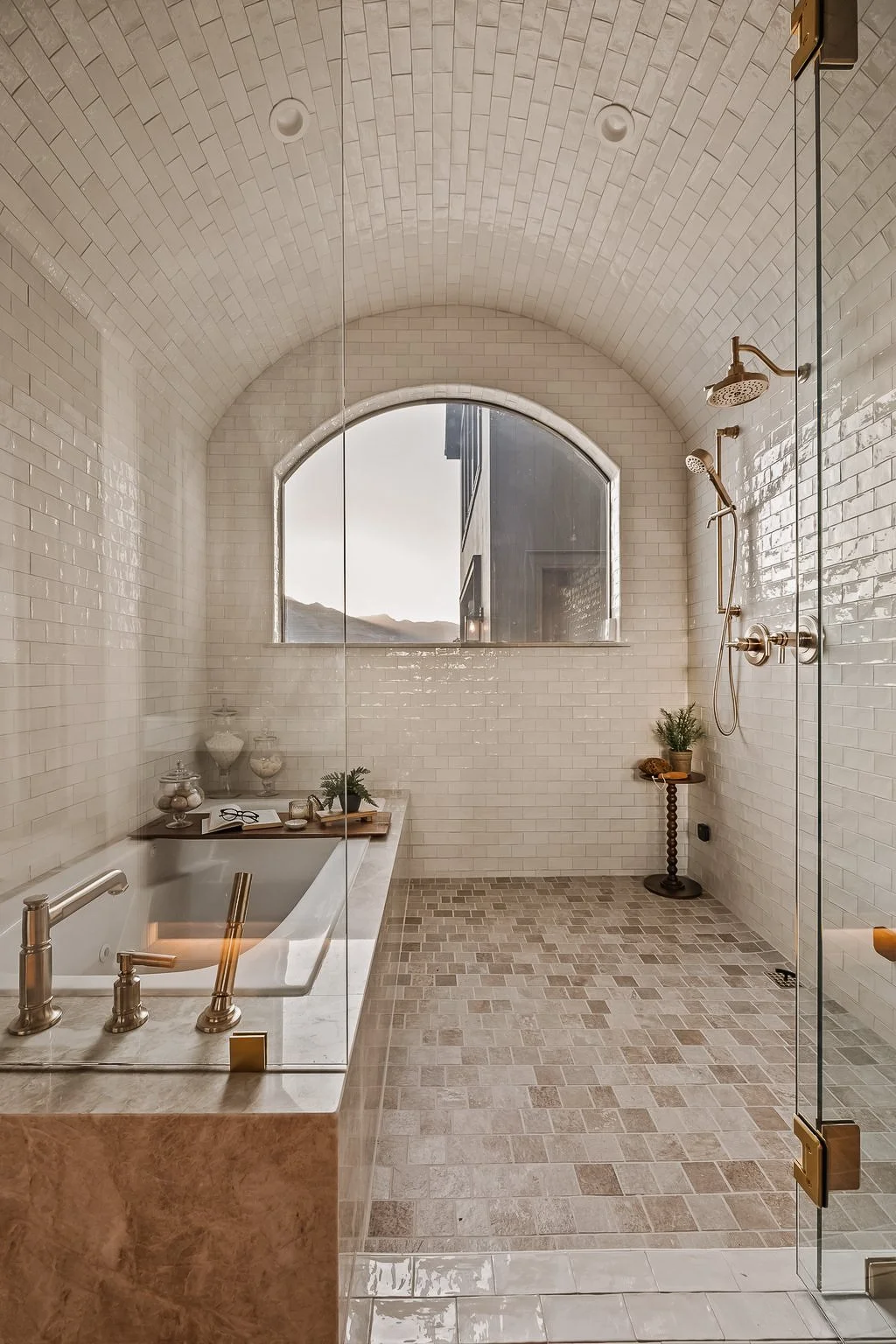 Modern bathroom with a large arched window, white subway tiles, a built-in bathtub with a marble surround, and a walk-in shower with gold fixtures. Decor includes potted plants, a small table, and decorative jars.