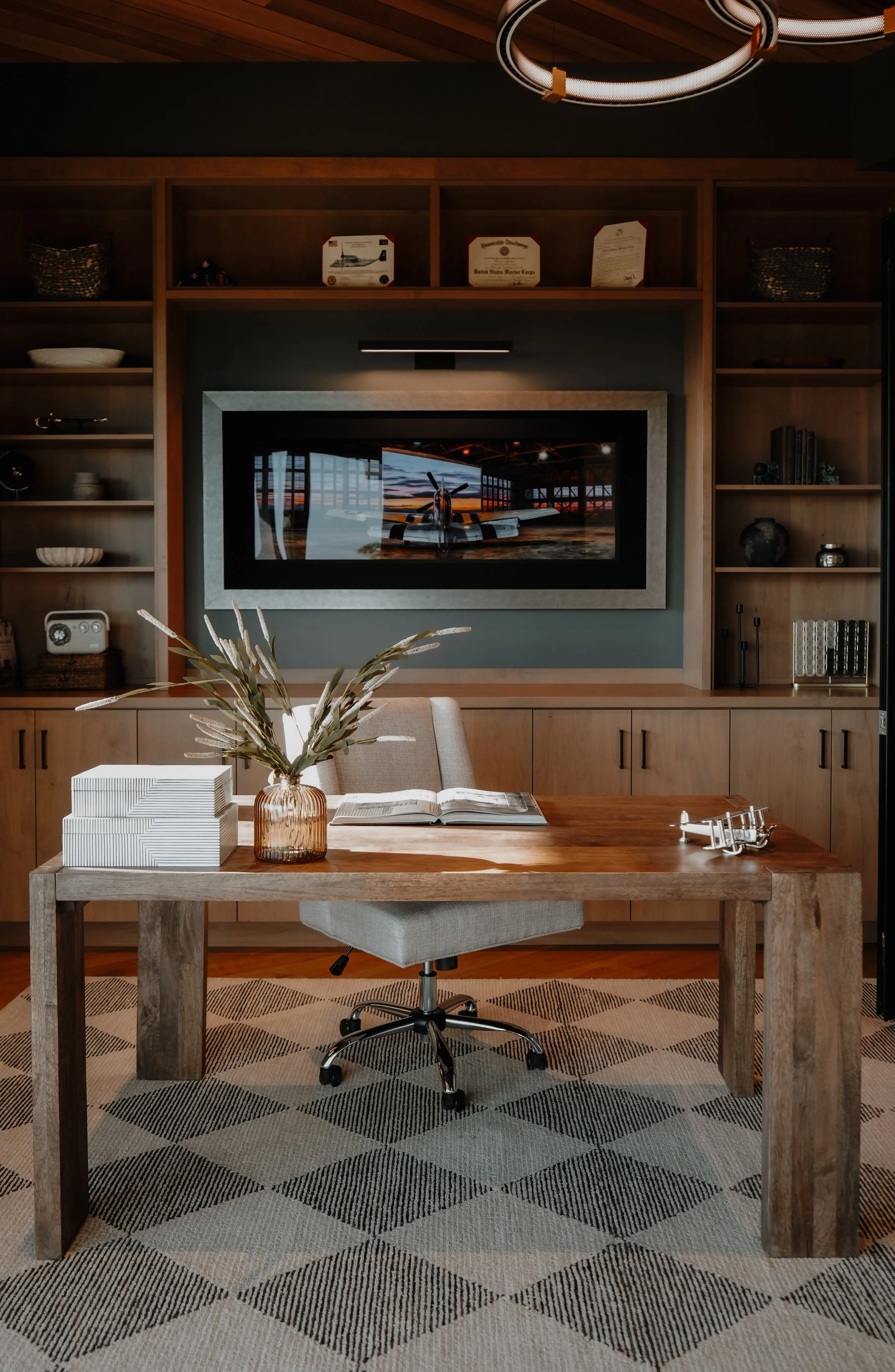 Home office with a wooden desk, an open book, a vase of leaves, and stacked books, set in front of a built-in shelves and a television displaying an aircraft photo.